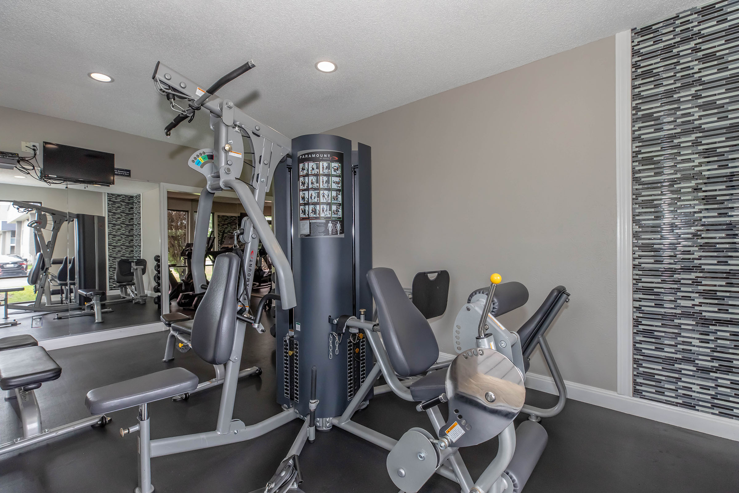 A modern gym interior featuring workout equipment, including multi-station strength machines and a bench. The room has neutral-colored walls and a stylish tiled accent wall, with natural light coming in from windows. A large television is mounted on the wall for entertainment during workouts.