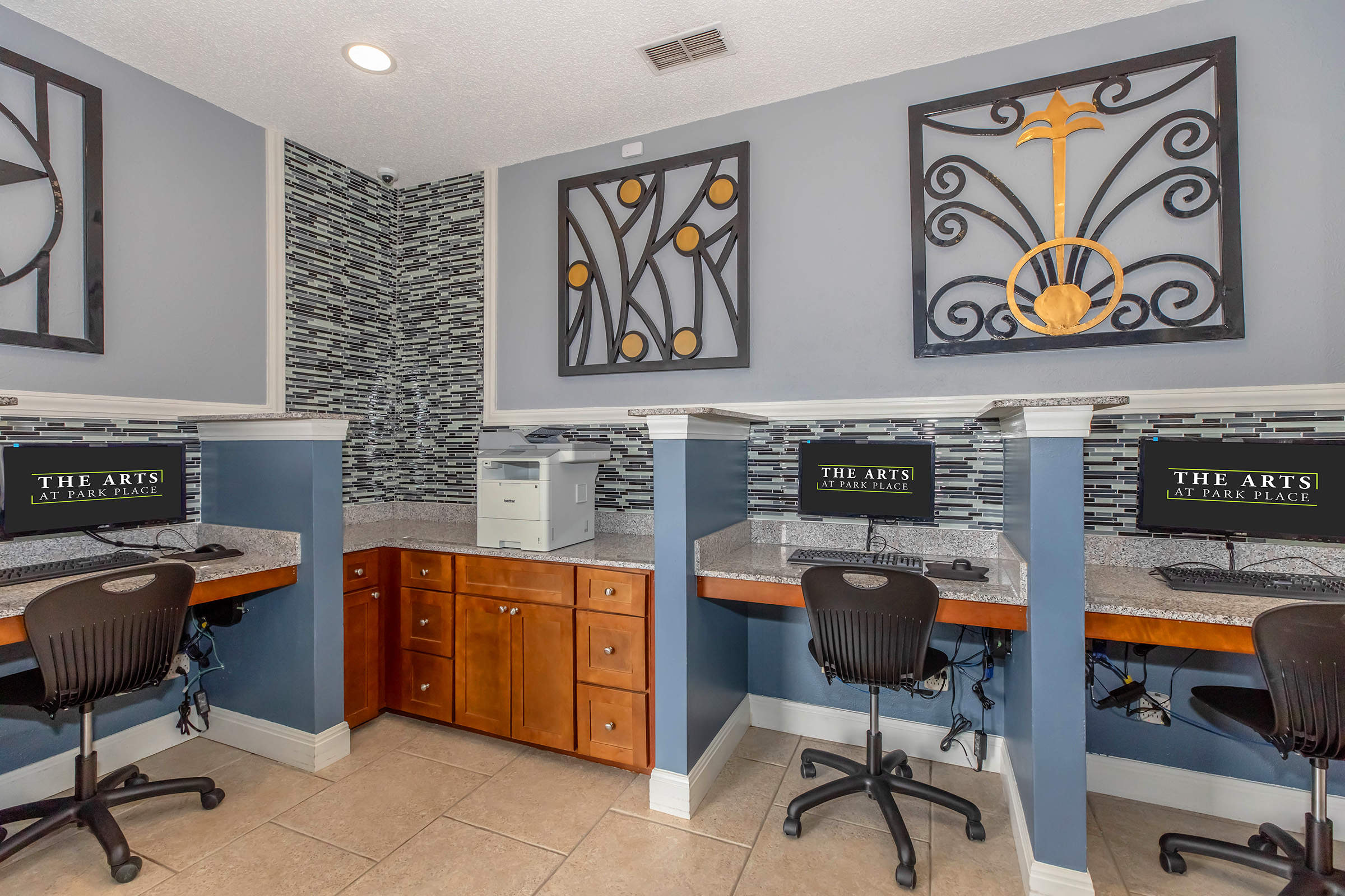 A modern workspace featuring two computer stations with black chairs. The walls are decorated with decorative metal art, and there is a printer on a cabinet nearby. The overall color scheme includes blue and gray tones, with a patterned tile backsplash.