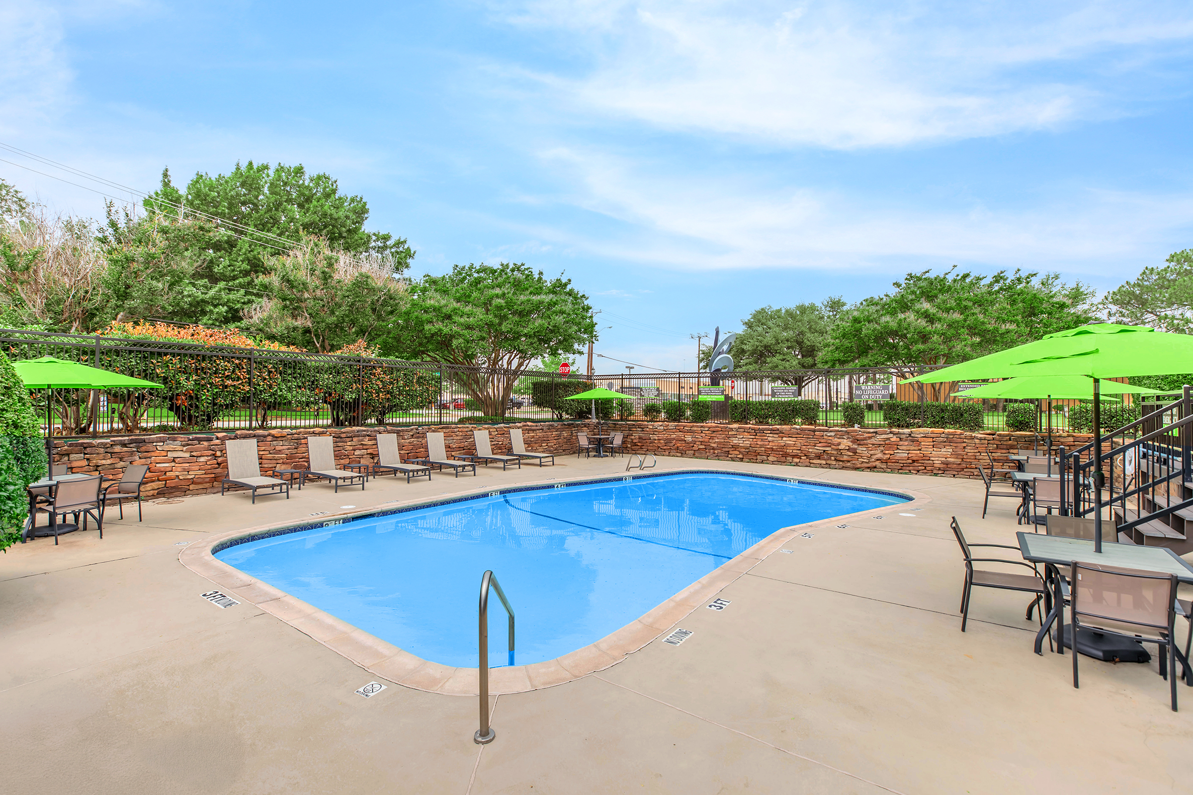 A serene outdoor pool area featuring a blue swimming pool surrounded by a stone wall. Sun loungers and patio tables with green umbrellas are arranged around the pool, set against a backdrop of lush trees and clear skies. Ideal for relaxation and leisure.