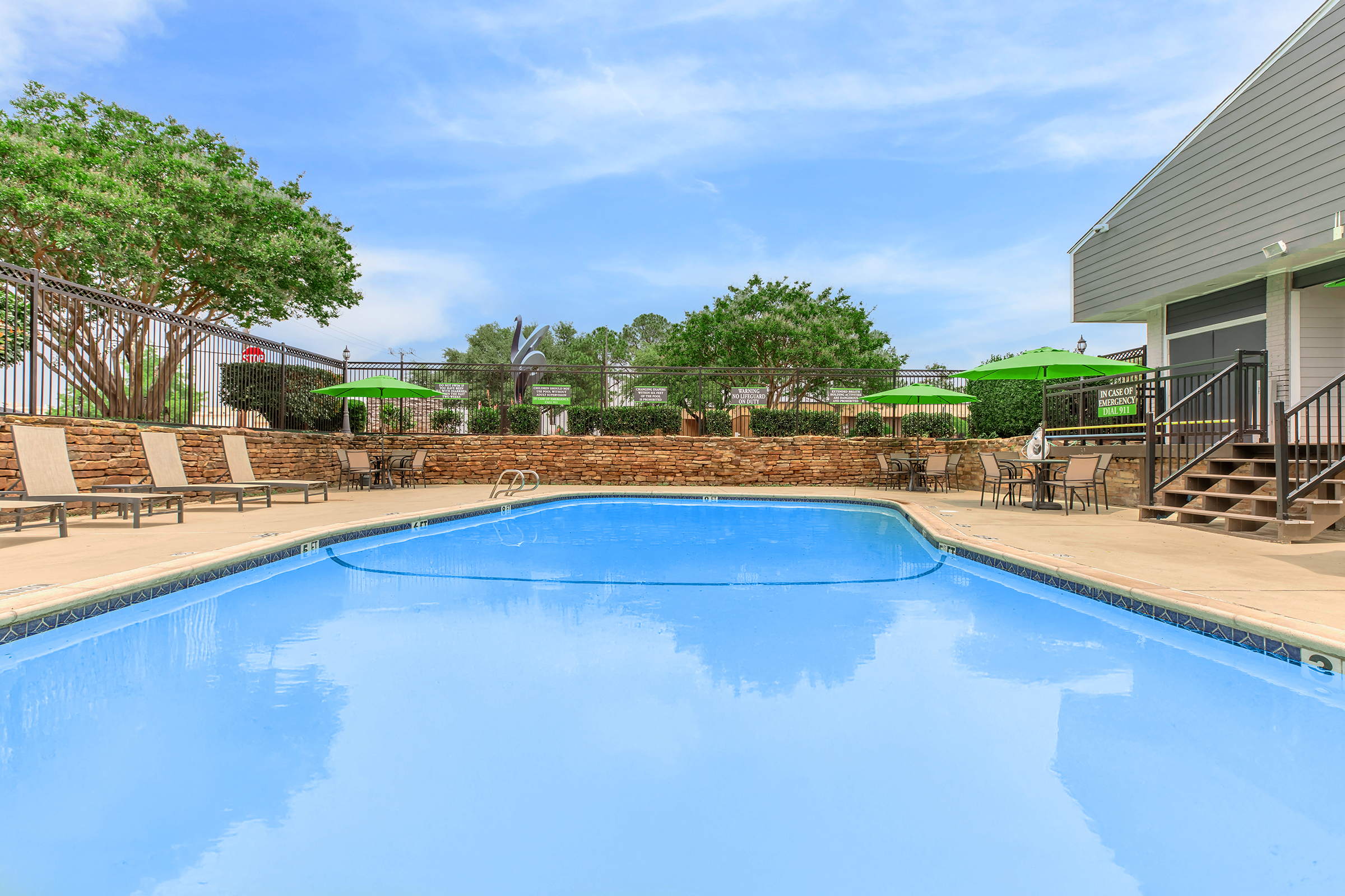 A clear blue swimming pool surrounded by lounge chairs on a patio. Green umbrellas provide shade in the background, along with lush greenery and a stone wall. The sky is bright with a few clouds, creating a serene outdoor space for relaxation.