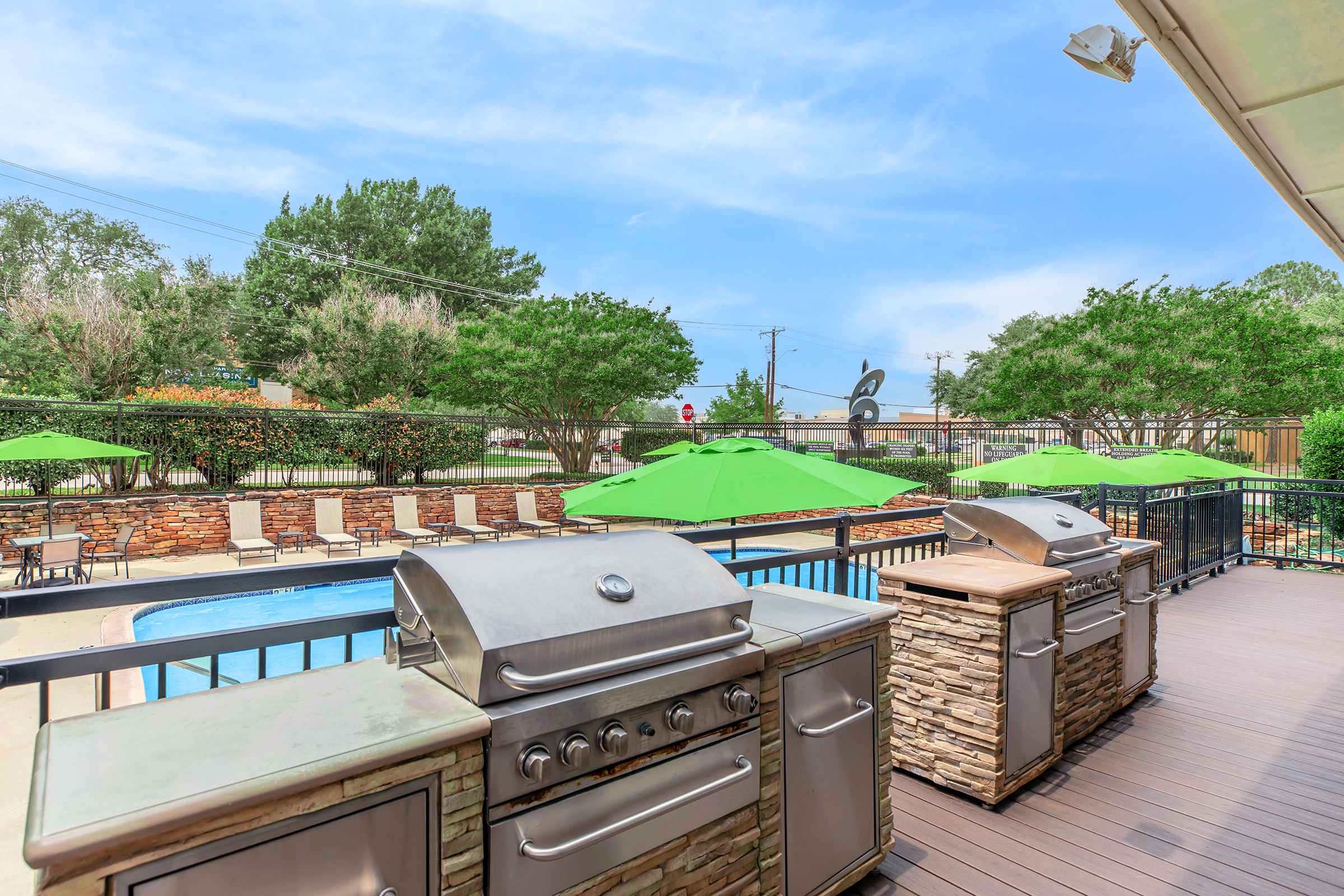 Outdoor grilling area featuring stainless steel barbecue grills on a wooden deck, overlooking a swimming pool. Bright green umbrellas provide shade for nearby seating areas, surrounded by lush landscaping and trees under a clear blue sky. Perfect for social gatherings and relaxation.