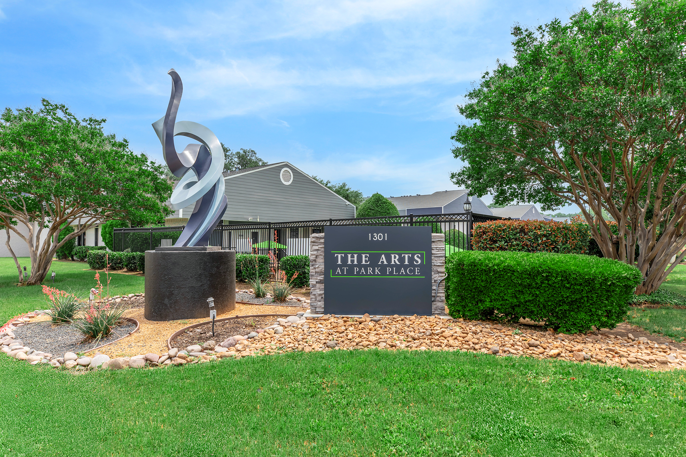 Sign for "The Arts at Park Place" with a modern sculpture in front, surrounded by landscaping featuring green grass, bushes, and decorative stones. The background includes a building, indicating a well-maintained property. The sky is clear with some clouds.
