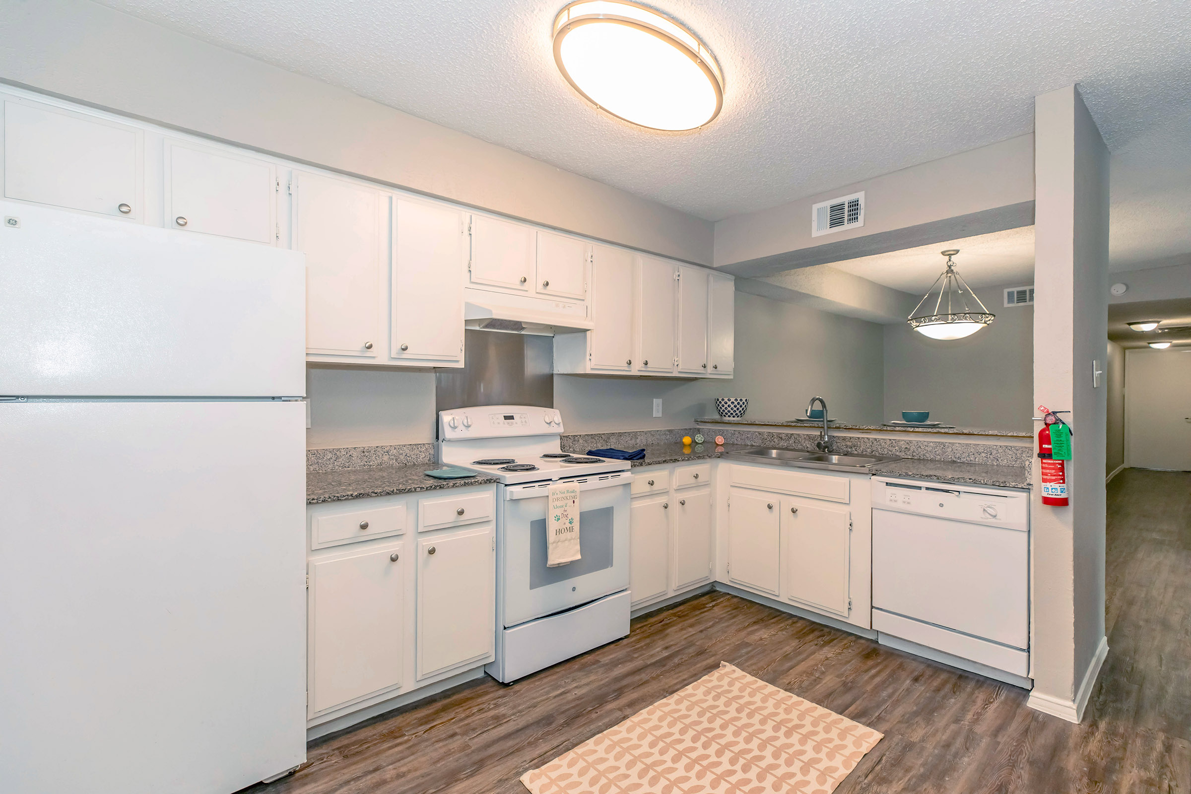Bright kitchen featuring white cabinets, a granite countertop, a white refrigerator, a stove with an oven, and a dishwasher. The space is well-lit with a circular ceiling light, has a decorative rug on the floor, and includes a view of a hallway leading to another room.