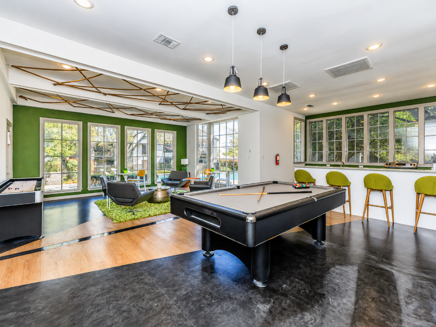 A modern game room featuring a black pool table in the foreground, surrounded by stylish seating. Large windows let in natural light, showcasing green walls and a contemporary design. Comfortable bar stools are positioned near a counter, adding to the inviting atmosphere of the space.