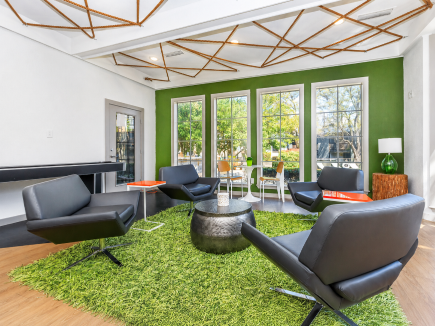 Modern lounge area featuring four black chairs arranged around a circular metallic table on a green carpet. Large windows let in natural light, showcasing a view of trees outside. The walls are painted in a lively green, with stylish lighting fixtures and a decorative ceiling design.