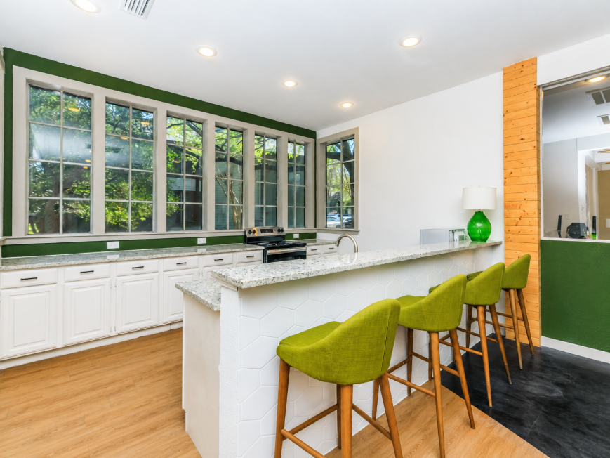A modern kitchen featuring green walls and a light wood accent wall. The space includes a countertop with three green bar stools, white cabinets, and large windows allowing natural light. A granite countertop and fixtures add a contemporary touch to the design.