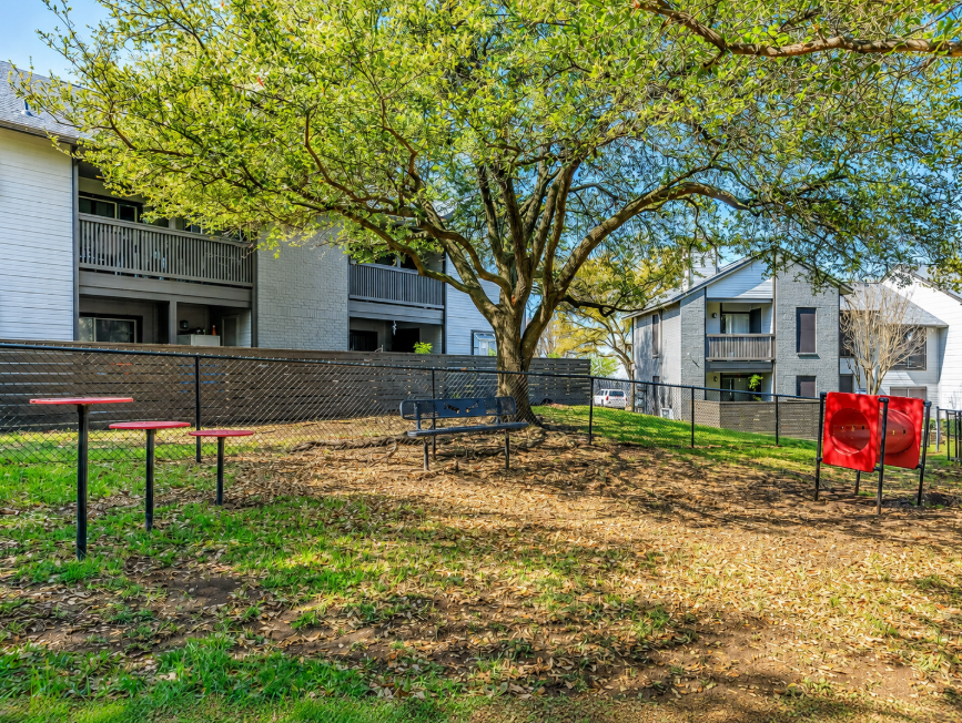 A grassy area with a large tree in the center, surrounded by several residential buildings. Beneath the tree, there’s a blue bench and red picnic tables. The scene is well-lit, with fresh green leaves and a fence in the background, creating a serene outdoor setting.