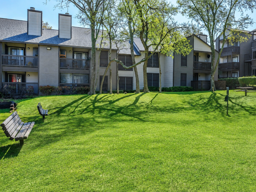 A well-maintained green lawn features several wooden benches in the foreground. In the background, there are multiple two-story residential buildings, each with balconies. Lush trees surround the area, casting shadows on the grass, creating a peaceful and inviting outdoor space.