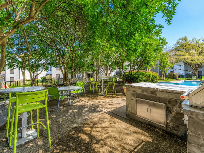 Outdoor patio area with green trees and shrubs, featuring several high-top tables and chairs in bright green. A grill is situated near a stone structure, and a swimming pool can be seen in the background. The scene is sunny, creating a relaxed atmosphere for gatherings.
