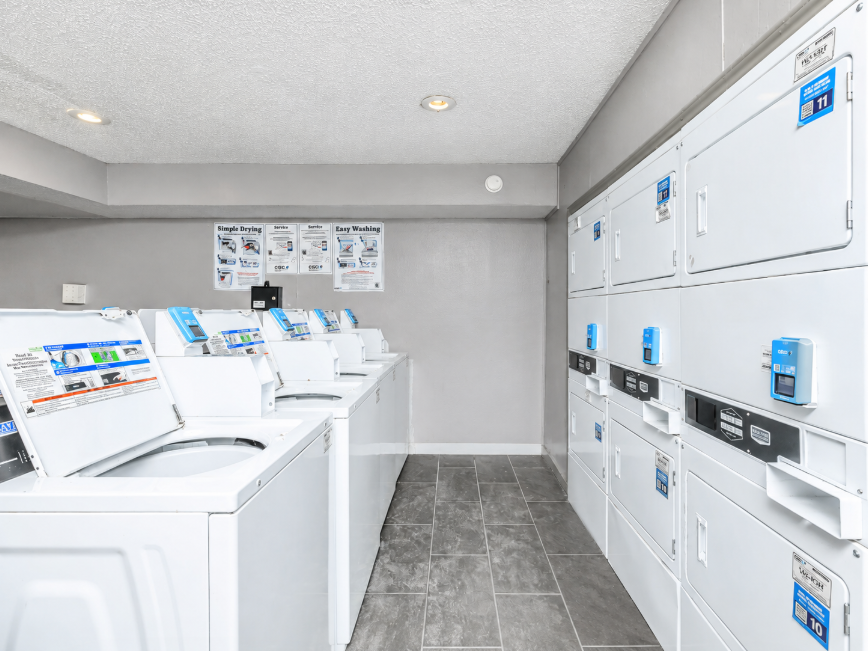 A clean and organized laundry room featuring several white washing machines lined up against the wall. The floor is tiled with gray tiles, and there are instruction signs and price lists mounted on the wall above the machines. The room is well-lit and has a modern design.