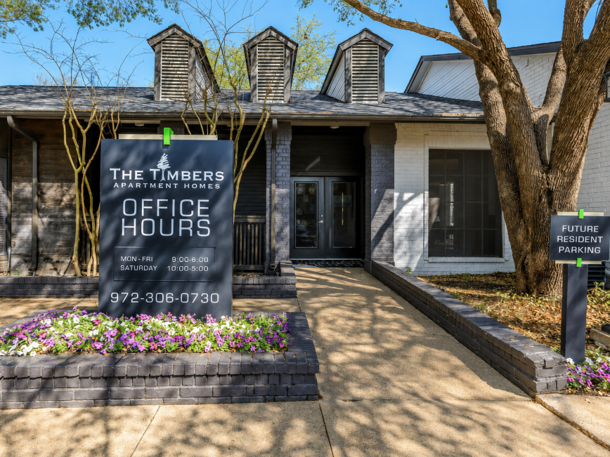 Exterior view of The Timbers Apartment Homes, featuring a sign that displays office hours: Monday to Friday, 9:00 AM - 6:00 PM and Saturday, 10:00 AM - 5:00 PM. There are flower beds in front and a sign indicating future resident parking. The building has a modern design with a dark roof and large windows.