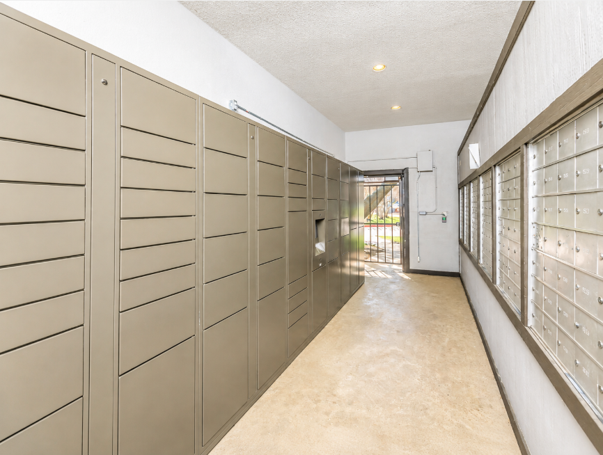 A corridor featuring metal mail lockers on the left side and wall-mounted mailboxes on the right. The space has a neutral color scheme with concrete flooring and natural light coming through windows. The area appears organized and well-lit, designed for secure mail delivery.