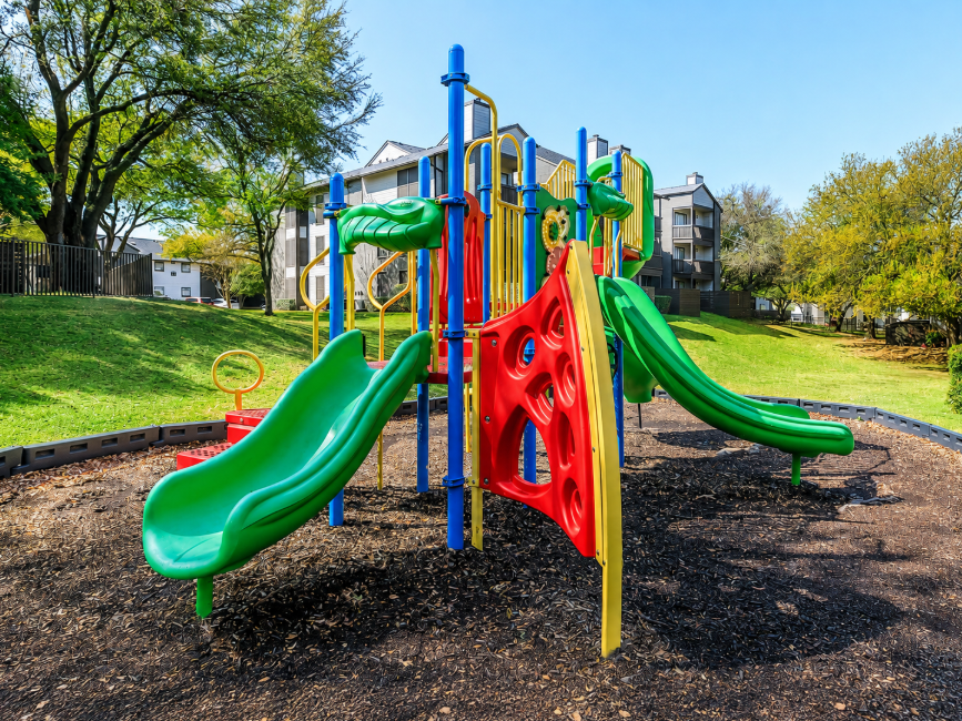 Colorful playground equipment featuring two green slides, a red climbing structure, and various play elements, set on a soft ground surrounded by grassy areas and trees. In the background, there are apartment buildings under a clear blue sky.