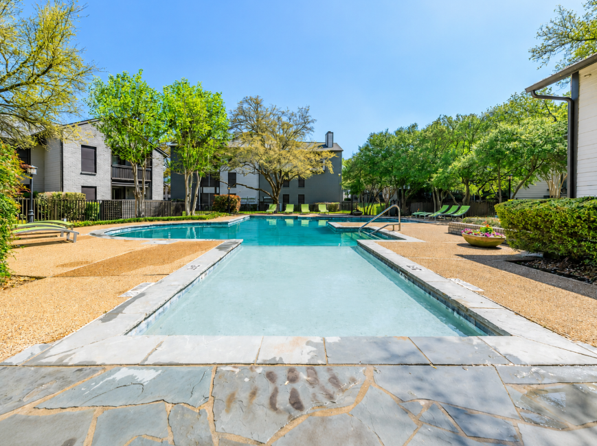 A clear, inviting swimming pool surrounded by a landscaped area featuring trees and shrubs. Sun loungers are positioned nearby, and the background includes several modern apartment buildings. The sky is bright blue, creating a sunny, relaxing atmosphere.