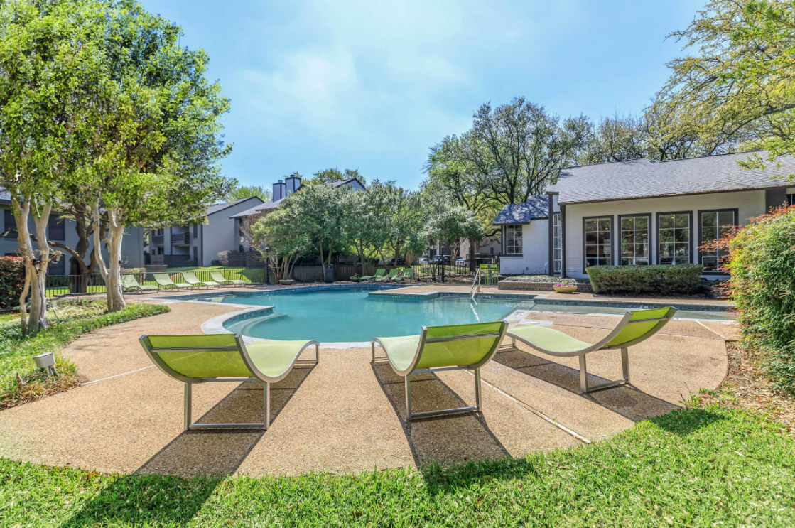 A serene pool area featuring a clear blue pool surrounded by green lounge chairs. Lush trees and well-maintained landscaping create a relaxing atmosphere, with residential buildings visible in the background under a bright blue sky. Ideal for leisure and relaxation.