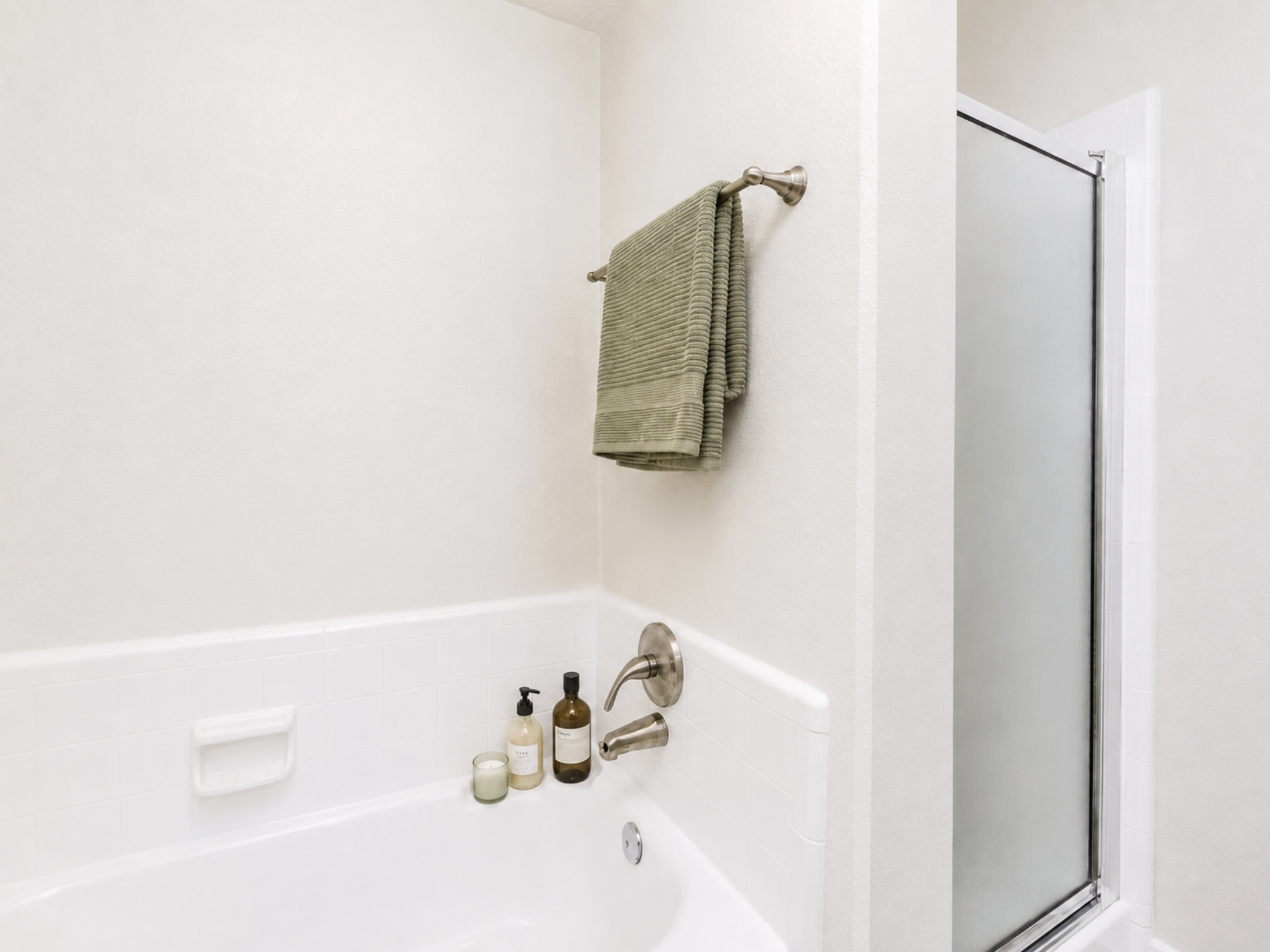 A clean bathroom corner featuring a white bathtub with a chrome faucet, a green towel hanging on a towel bar, and shower products on a ledge. The wall is light-colored, and there's a frosted glass shower door on the right side.