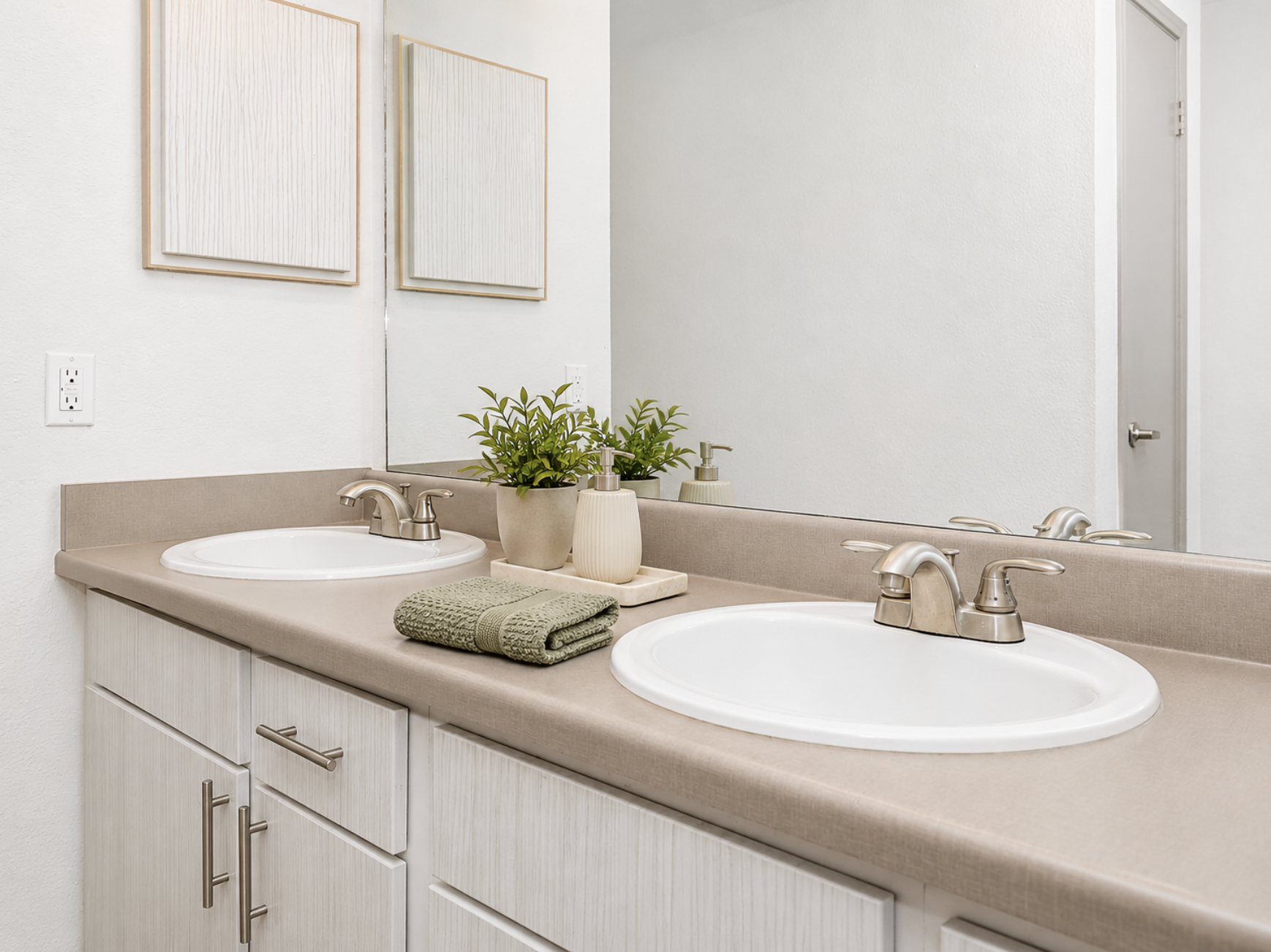 A modern bathroom featuring a double sink with silver faucets, light-colored cabinetry, and a neutral countertop. Above the sinks are two framed mirrors and a small green plant in a ceramic pot. A neatly rolled green towel is placed next to a decorative tray on the countertop. The overall ambiance is clean and minimalist.