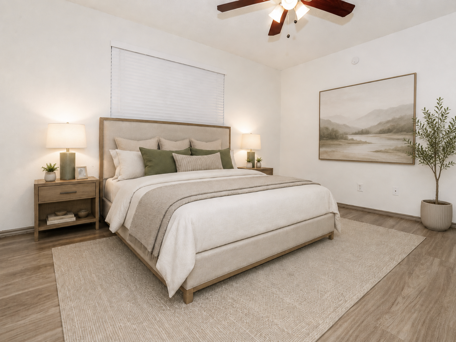 A cozy bedroom featuring a large bed with a light-colored blanket and decorative pillows. Two bedside tables with lamps flank the bed. A neutral area rug covers the floor, and a framed landscape painting hangs on the wall. A small indoor plant adds a touch of greenery, while a ceiling fan completes the serene atmosphere.