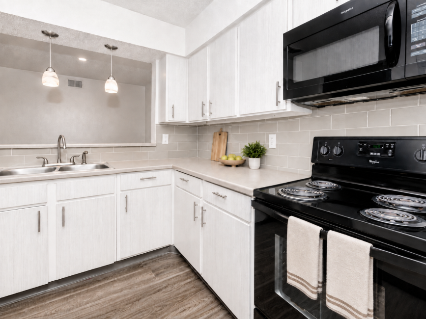 Modern kitchen featuring white cabinetry, a stainless steel sink, and black appliances including a microwave and stove. Countertops are light-colored with a wood grain floor. Two pendant lights hang above the sink, and decorative items like a cutting board and a bowl of limes are on the counter.