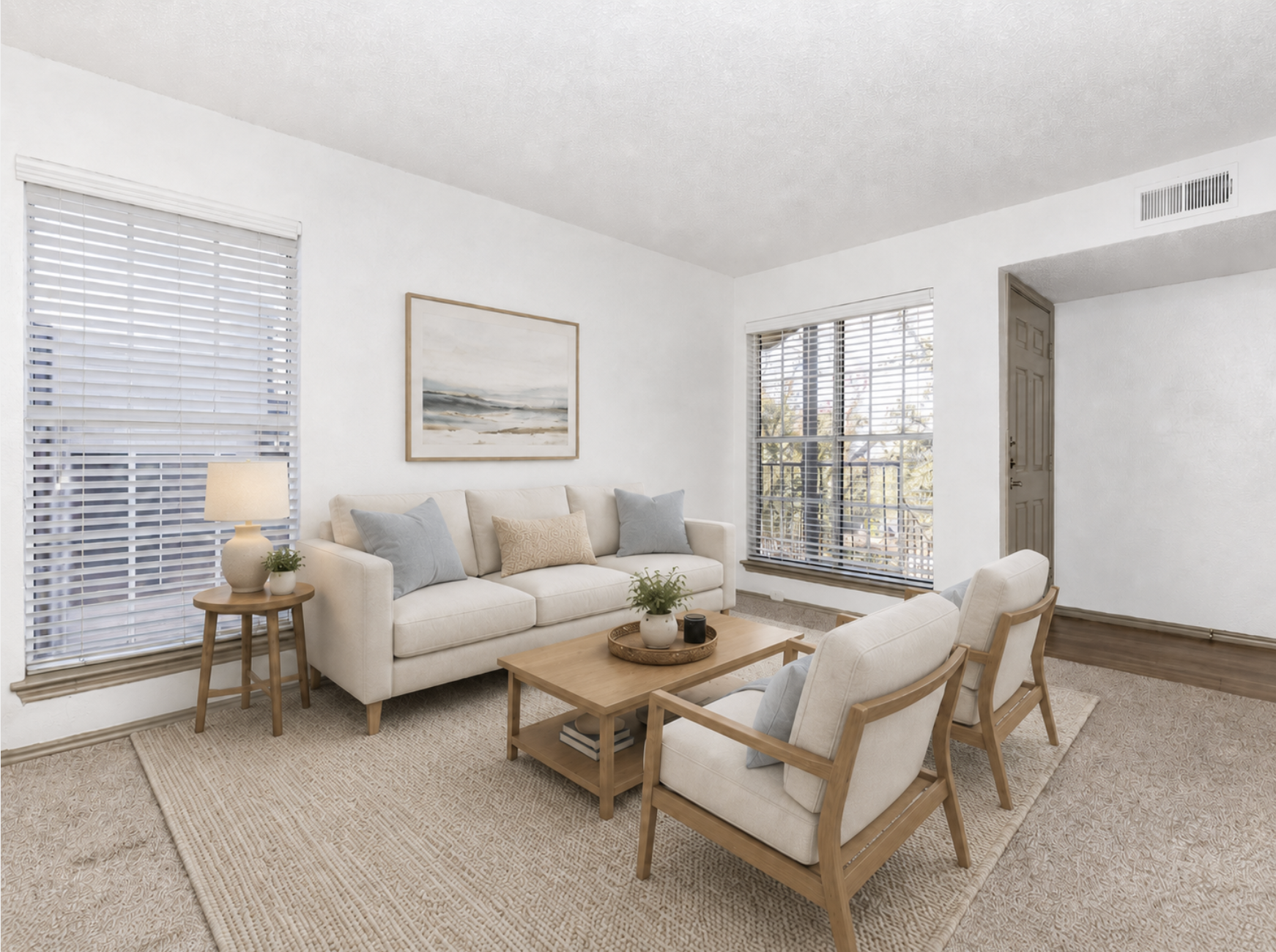A bright and cozy living room featuring a light-colored sofa with decorative pillows, two wooden armchairs, a round coffee table, and a lamp on a side table. Large windows with blinds allow natural light, and a simple landscape painting adorns the wall. A soft area rug enhances the inviting atmosphere.