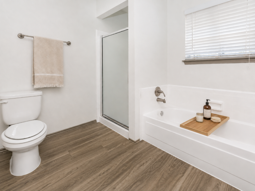 A modern bathroom featuring a white toilet, a glass shower, and a white bathtub with a wooden tray holding decorative bottles. The walls are light-colored, and there is a towel hanging on a rail. Natural light enters through a small window with blinds. The flooring is wood-look laminate.