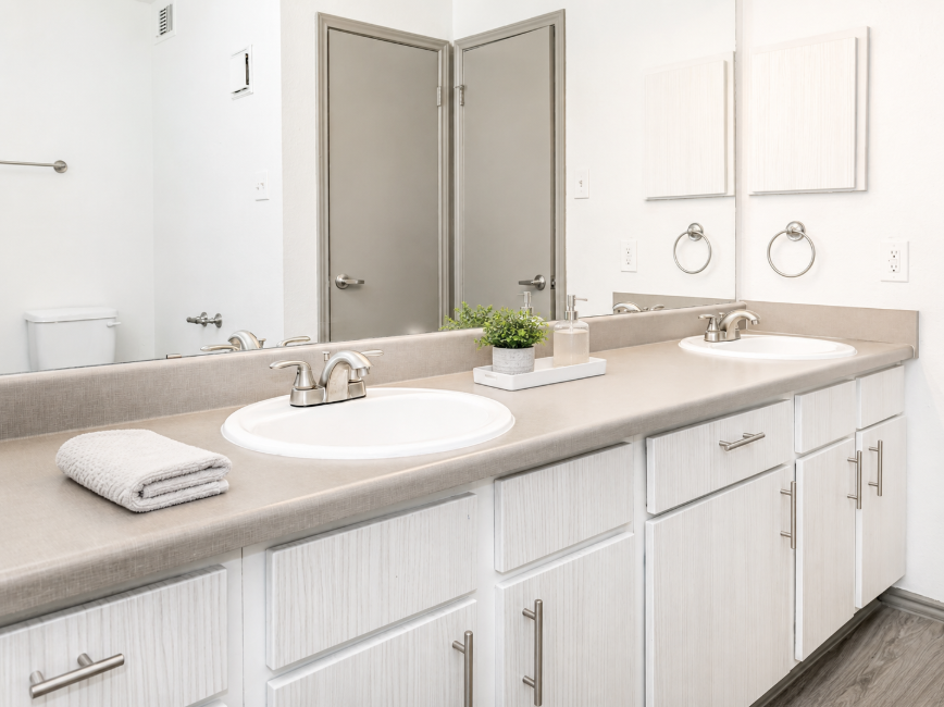 Bright and modern bathroom featuring double sinks with a light gray countertop. The cabinetry is white with horizontal grooves. A decorative plant and candle are placed between the sinks. A towel is neatly rolled and placed nearby. Mirrors above the sinks reflect the clean, minimalist design.
