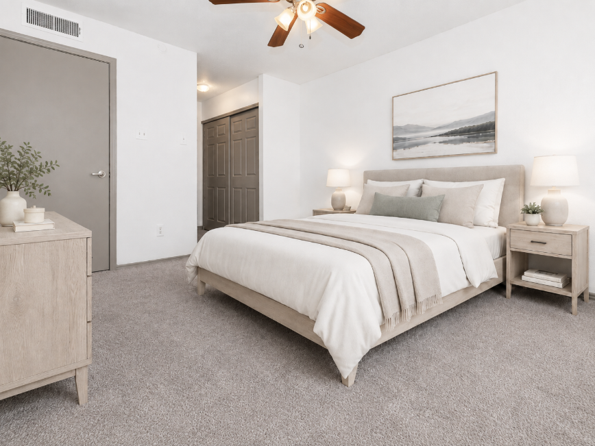 A cozy and modern bedroom featuring a light wood bed frame with a neutral-colored bedspread and pillows. There are matching nightstands with lamps on either side, and a piece of abstract wall art above the bed. The room has light-colored walls, carpeted flooring, and a ceiling fan, creating a warm and inviting atmosphere.