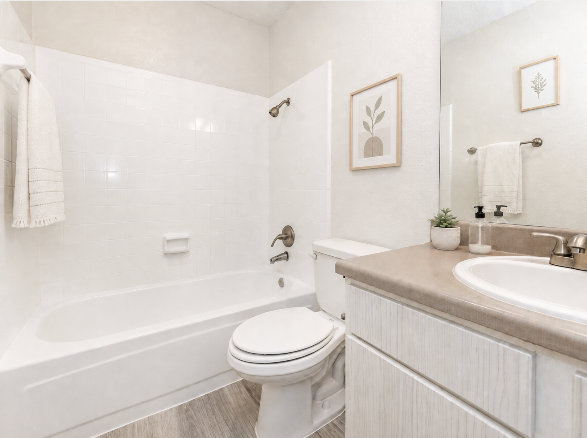 A modern bathroom featuring a white bathtub with a shower, a beige countertop with sinks, and a small plant. Light-colored walls and flooring enhance the bright, clean atmosphere. A decorative framed plant print is on the wall, and there are neatly arranged towels and toiletries.