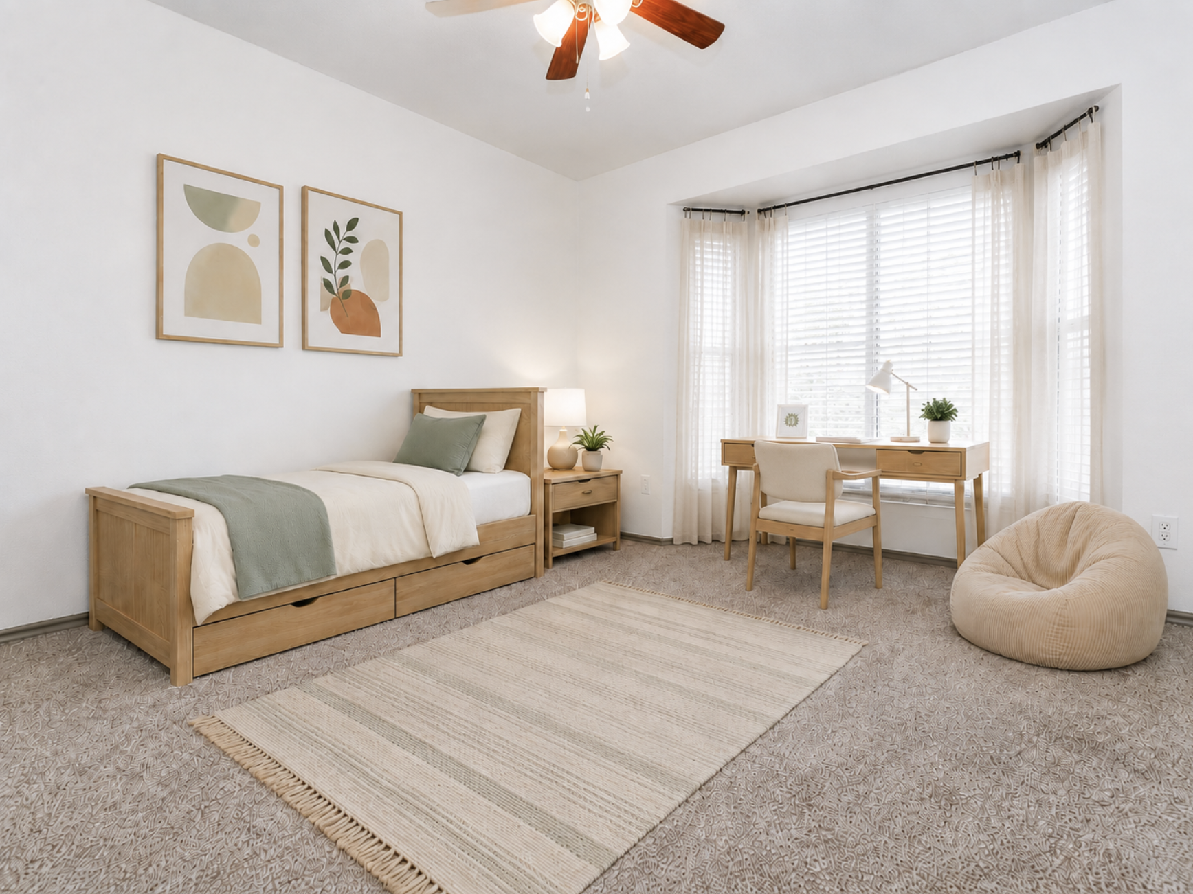 A cozy bedroom featuring a single bed with green and beige bedding, a small desk with a lamp and chair, and a beanbag. Two framed artworks hang on the wall, showcasing abstract shapes. The room has light-colored walls, large windows with sheer curtains, and a soft area rug on carpet flooring.