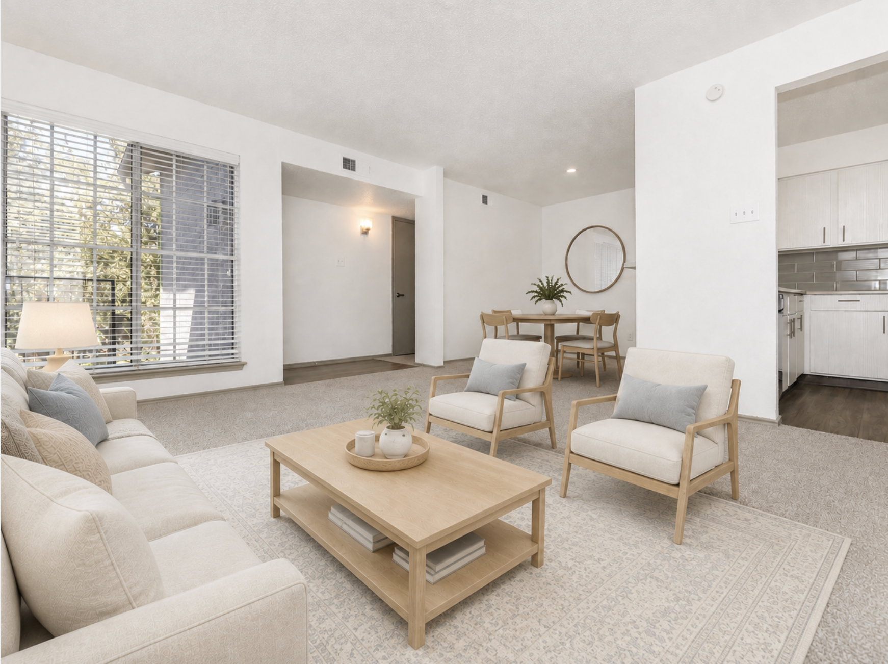 A modern living room featuring a light-colored sofa and two armchairs, a wooden coffee table with decor, and a large window with blinds allowing natural light. A dining area with a round table is visible in the background, and a kitchen can be seen to the right. The space has a neutral color palette and a cozy atmosphere.