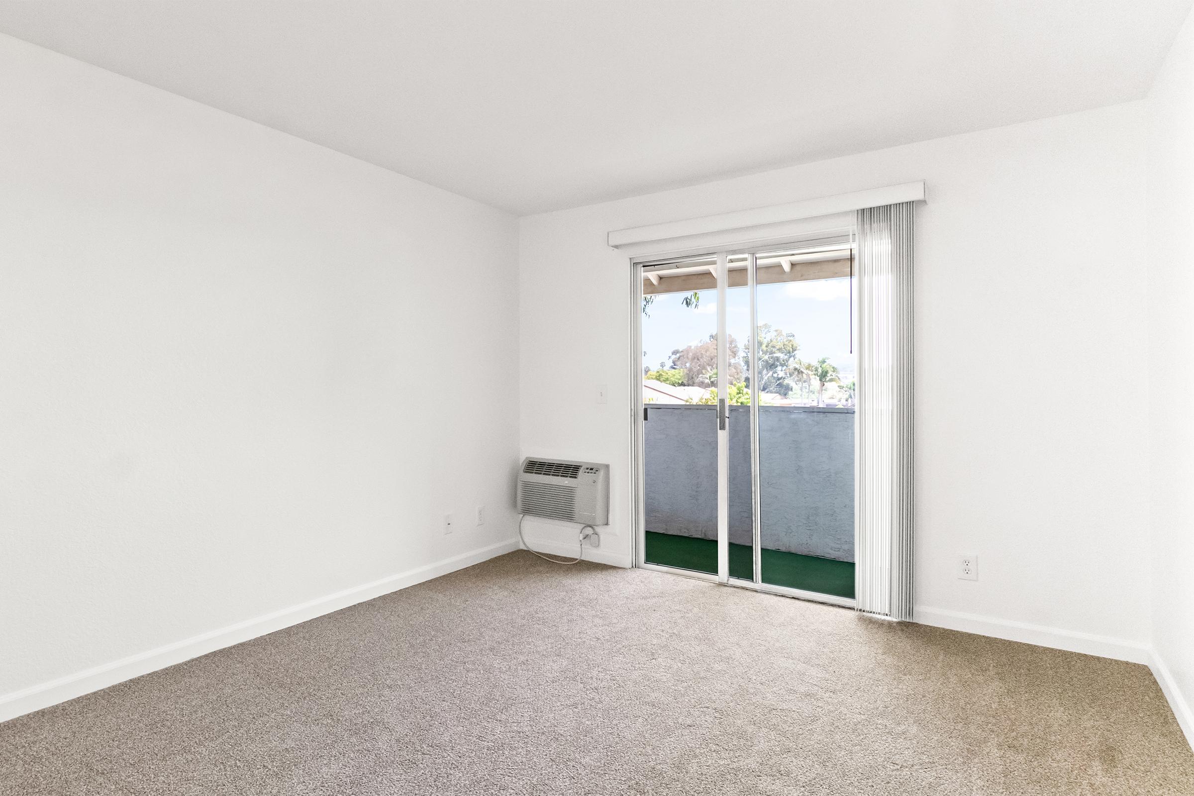 A sparsely furnished room featuring light-colored walls and a tan carpeted floor. The space includes a large sliding glass door leading to a balcony, with an air conditioning unit mounted on the wall. Natural light floods the room, creating an airy atmosphere.