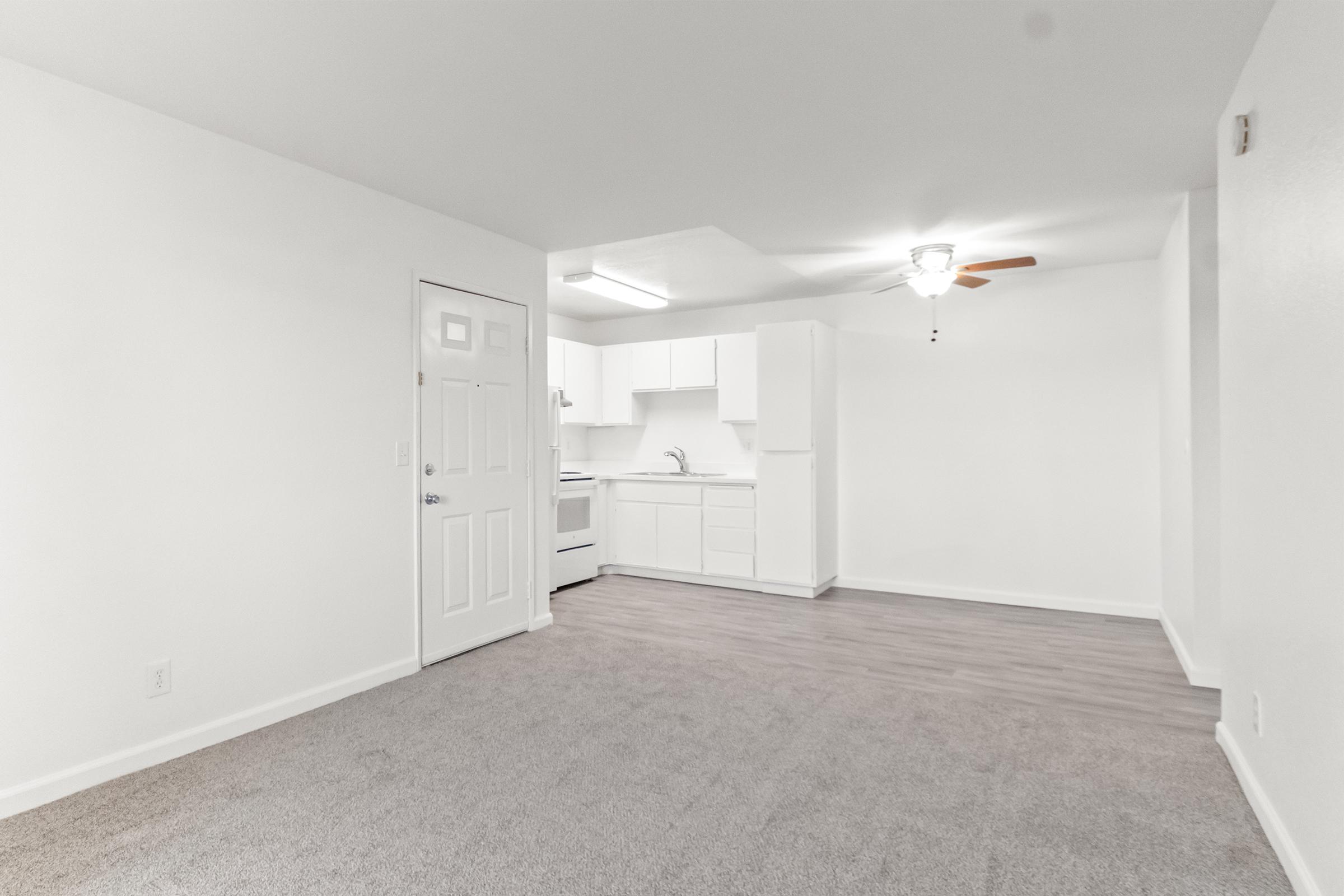 A bright, unfurnished living space featuring light gray carpet, a door leading to the hallway, and an open kitchen area with white cabinets, a stove, and a sink. The room is well-lit with a ceiling fan and has neutral white walls, creating a clean and modern look. Ideal for personal customization.