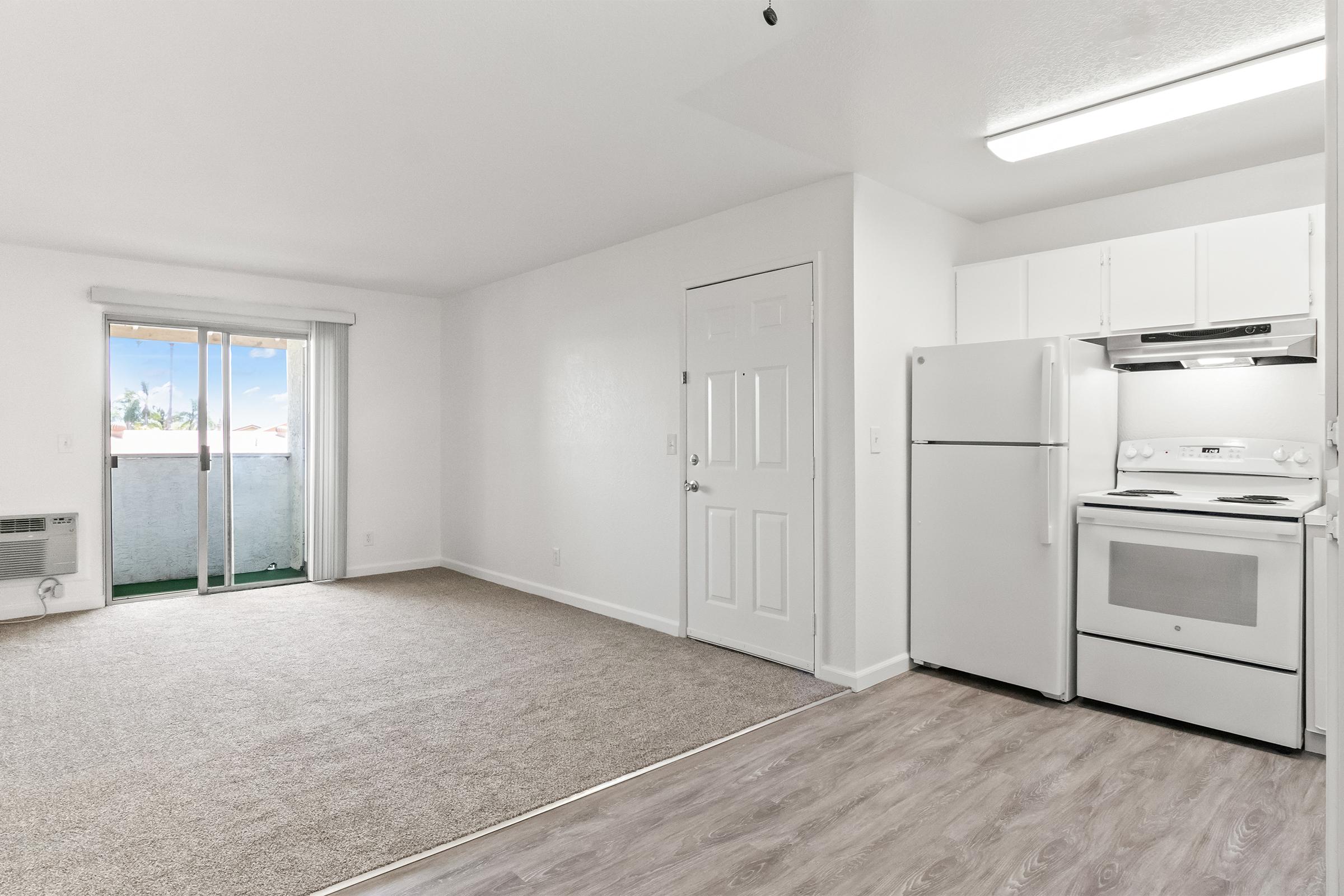 A bright and inviting living space featuring neutral-colored walls and carpet. The room includes a compact kitchen area with white appliances, including a stove and refrigerator. A sliding glass door leads to a small balcony, allowing natural light to enter the space.