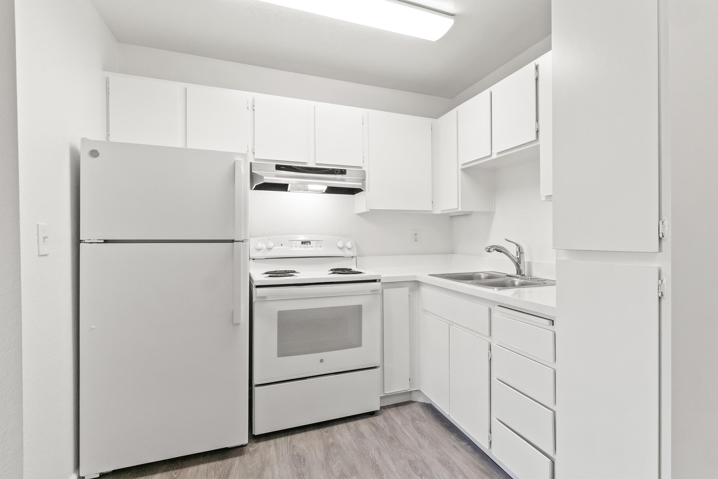 A modern, clean kitchen featuring white cabinetry, a refrigerator, an oven with a stovetop, and a sink. The walls are painted white, and the flooring is a light wood laminate. The kitchen has ample storage space with drawers and cabinets, creating a bright and airy atmosphere.