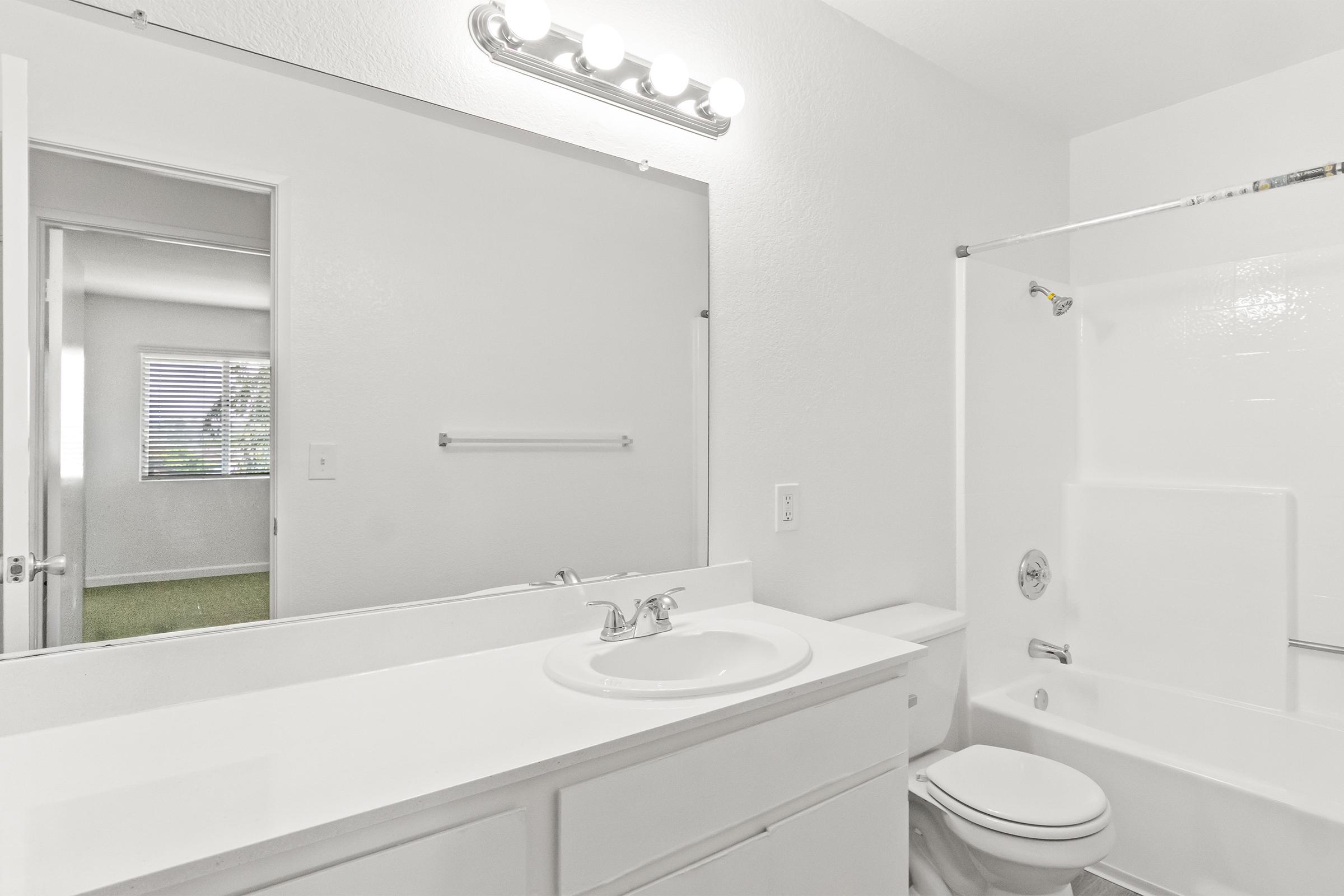 A bright and clean bathroom featuring a white countertop with a sink, a large mirror, and modern lighting above. There is a shower/tub combo, a toilet, and a towel rack. A door leads to another room, visible in the background, with a window showing greenery outside.