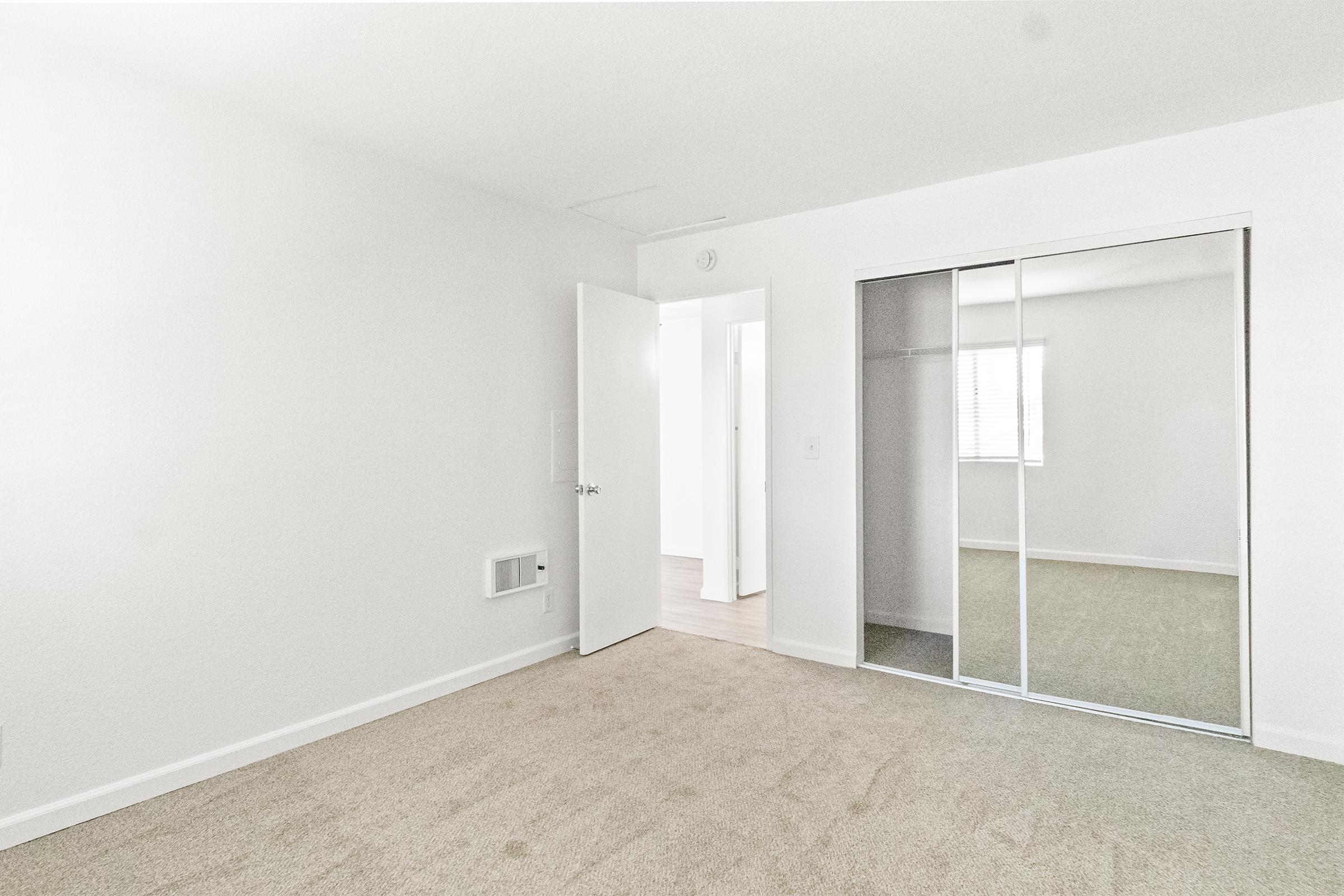 A spacious, empty room with light-colored walls and carpeted flooring. There is a white door partially open leading to another space, a large mirrored closet on the left, and a window with blinds. The overall atmosphere is bright and airy, creating a clean slate for decoration.