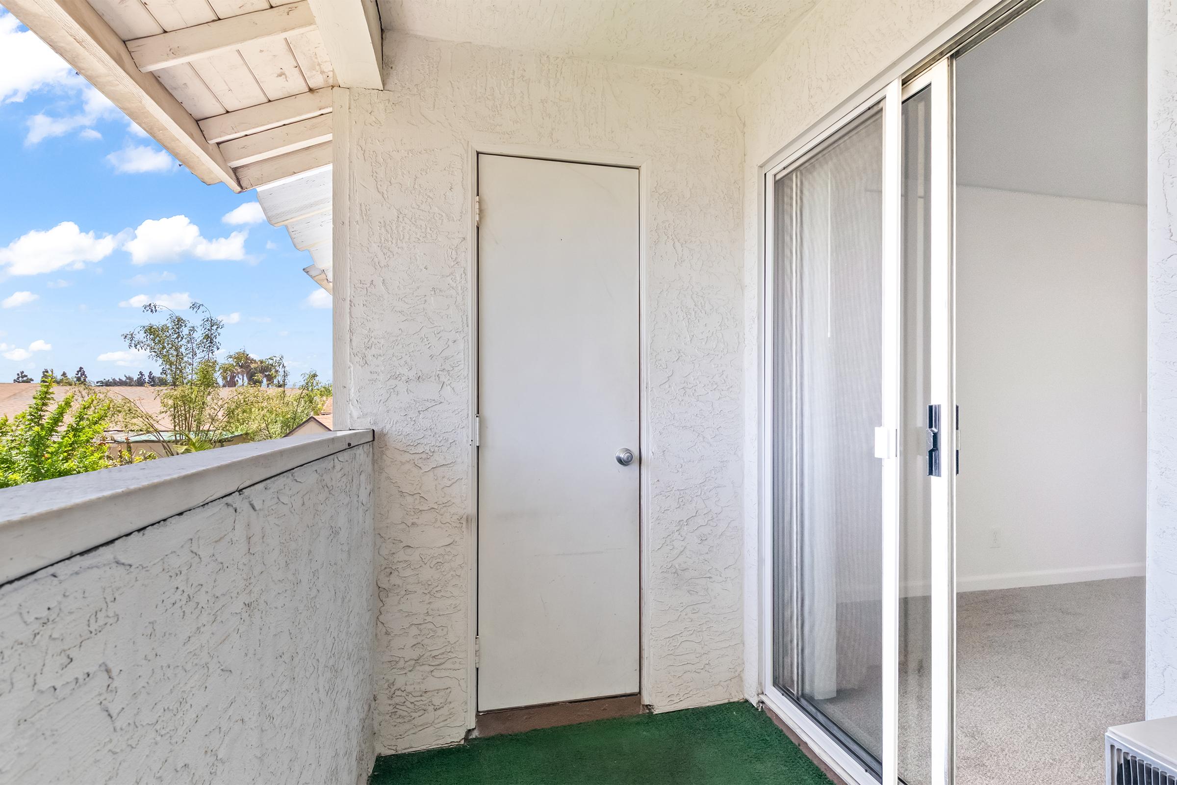 A balcony with a sliding glass door and a solid door. The balcony has textured walls and a green carpet, with a view of trees and a blue sky dotted with clouds in the background. The scene is bright and inviting, showcasing a residential outdoor space.