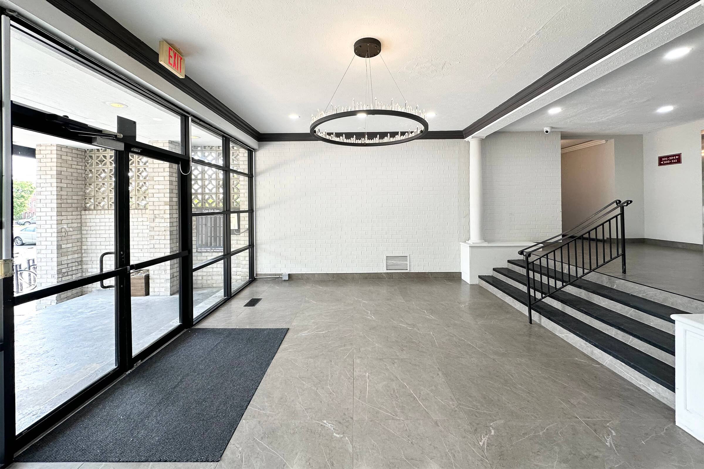 A well-lit entryway with a modern chandelier hanging from the ceiling. The floor is covered in gray tiles, and there is a large glass door leading outside. To the right, a set of stairs leads to another level, while the left side features an entrance mat. The walls are painted white, giving a clean look.