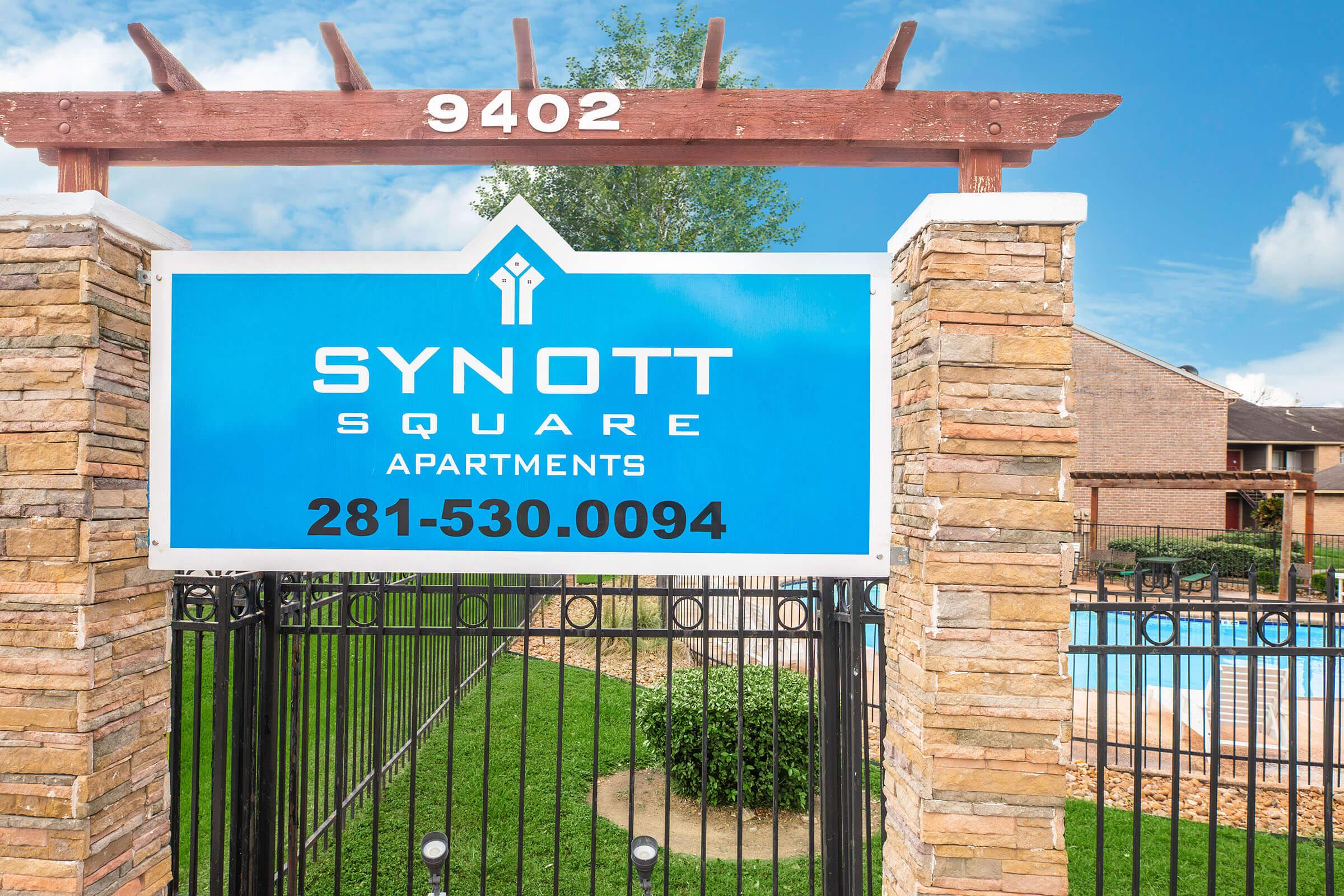 A blue sign reading "SYNOTT SQUARE APARTMENTS" with the phone number "281-530-0094," surrounded by stone pillars and a black gate. In the background, there's a glimpse of a swimming pool and green landscaping. The scene is under a clear blue sky.