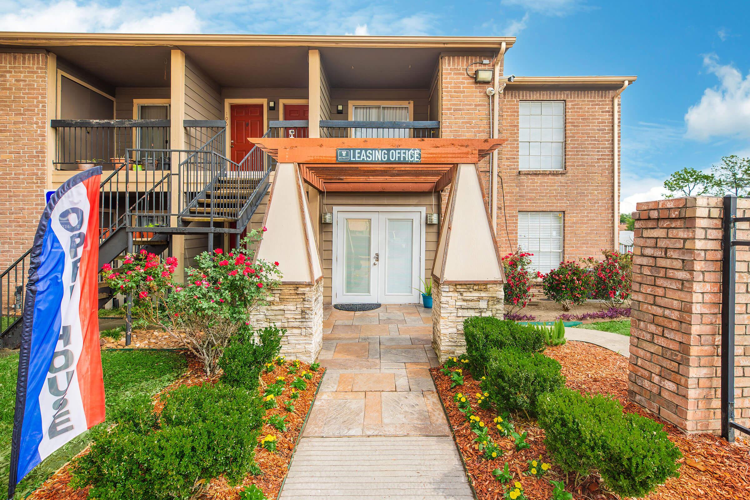 Exterior view of a leasing office entrance with a welcome sign. The building features multiple levels, surrounded by well-maintained landscaping including flowers and shrubs. A colorful flag with "OPEN" is displayed prominently, indicating the office is open for business. Clear sky in the background.