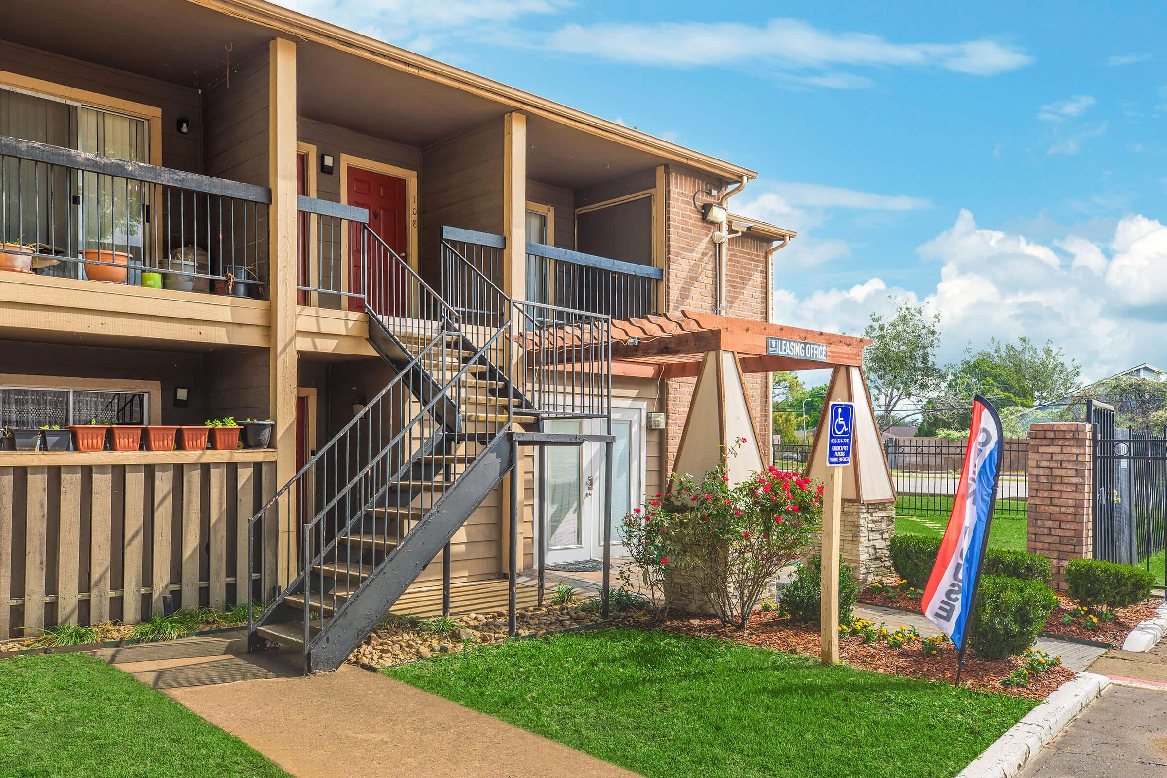 A two-story apartment building with a wooden staircase leading to the second floor. The entrance is adorned with potted plants, and there's a sign indicating a accessible parking space. Green grass and flowering shrubs surround the walkway, with a bright blue sky in the background.