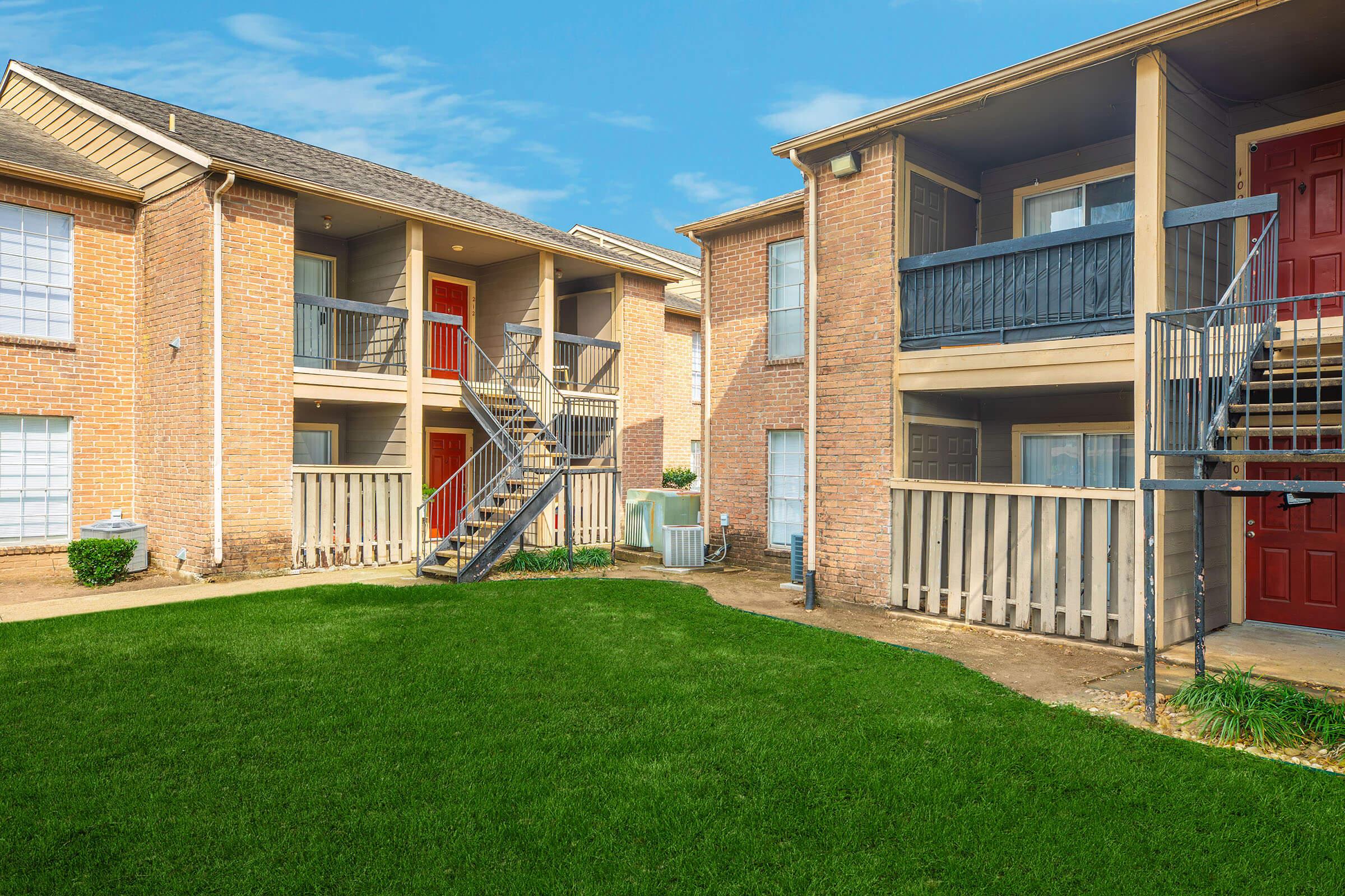 Two-story apartment complex with brick exterior. The building features a grassy area in front, and has outdoor stairs leading to upper units with balconies. Some apartments have red doors. Clear blue sky above and well-maintained landscaping.
