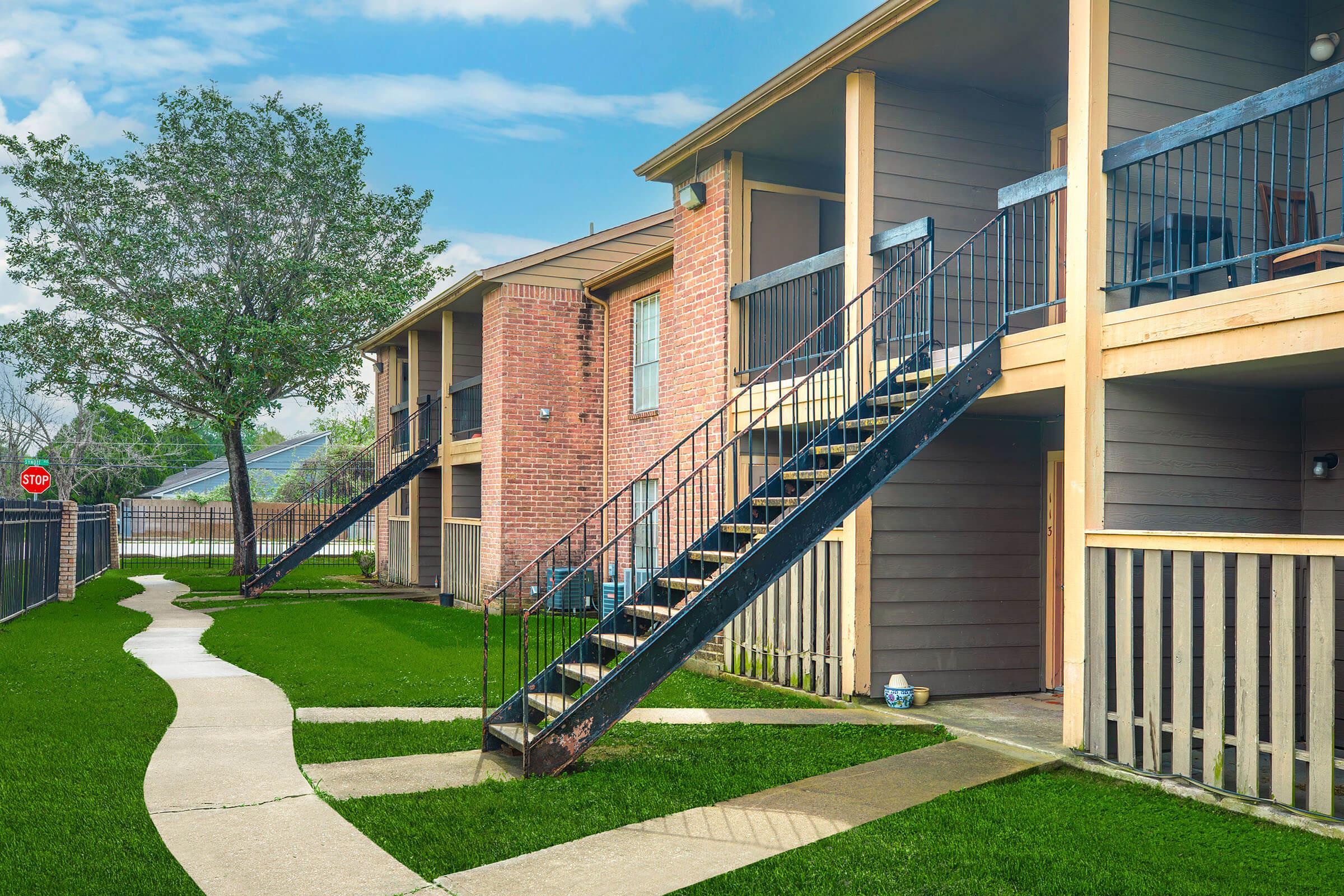 Exterior view of an apartment complex featuring two-story buildings with wooden balconies and black metal staircases. A winding concrete path leads through a well-maintained lawn, flanked by a tree. A stop sign is visible in the background. The sky is partly cloudy.