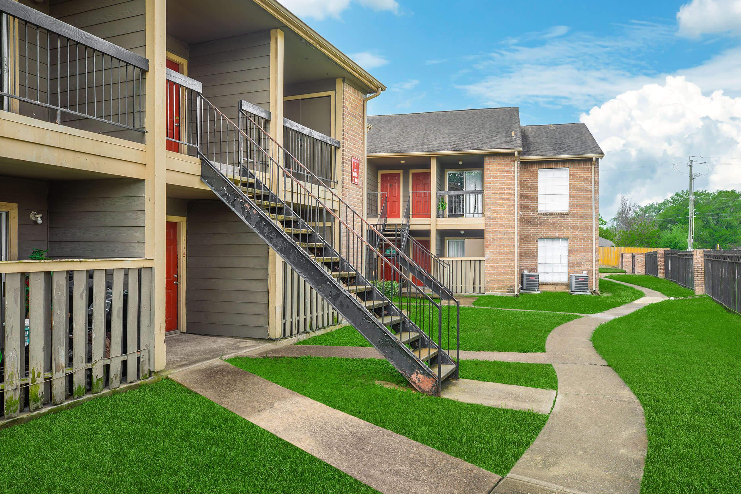 Two multi-story apartment buildings with a grassy area in front. A staircase leads to the upper level of one building, while a concrete pathway winds between the structures. The exterior features brick and wooden elements, with red doors visible. The sky is partly cloudy, indicating a clear day.
