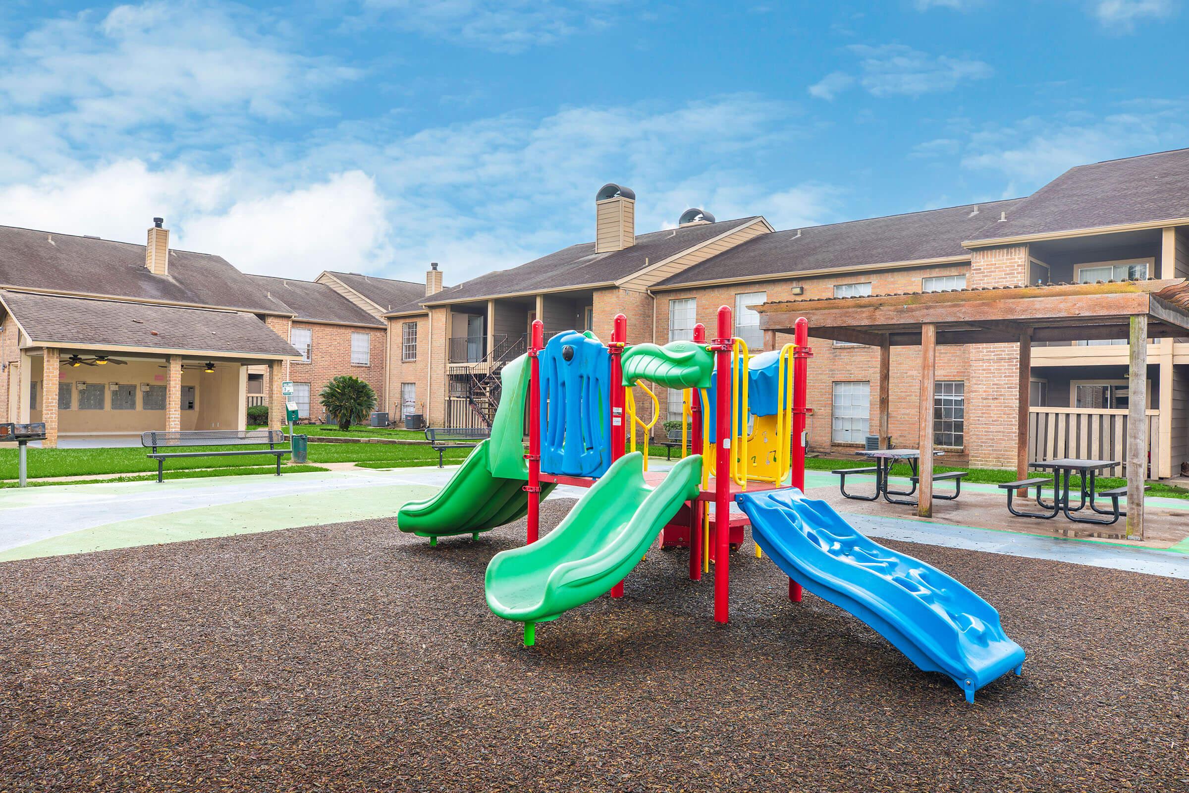 A colorful playground featuring blue and green slides, surrounded by gravel and grass. In the background, there are brick apartment buildings, benches, and a clear blue sky. The setting suggests a communal outdoor space for children to play.