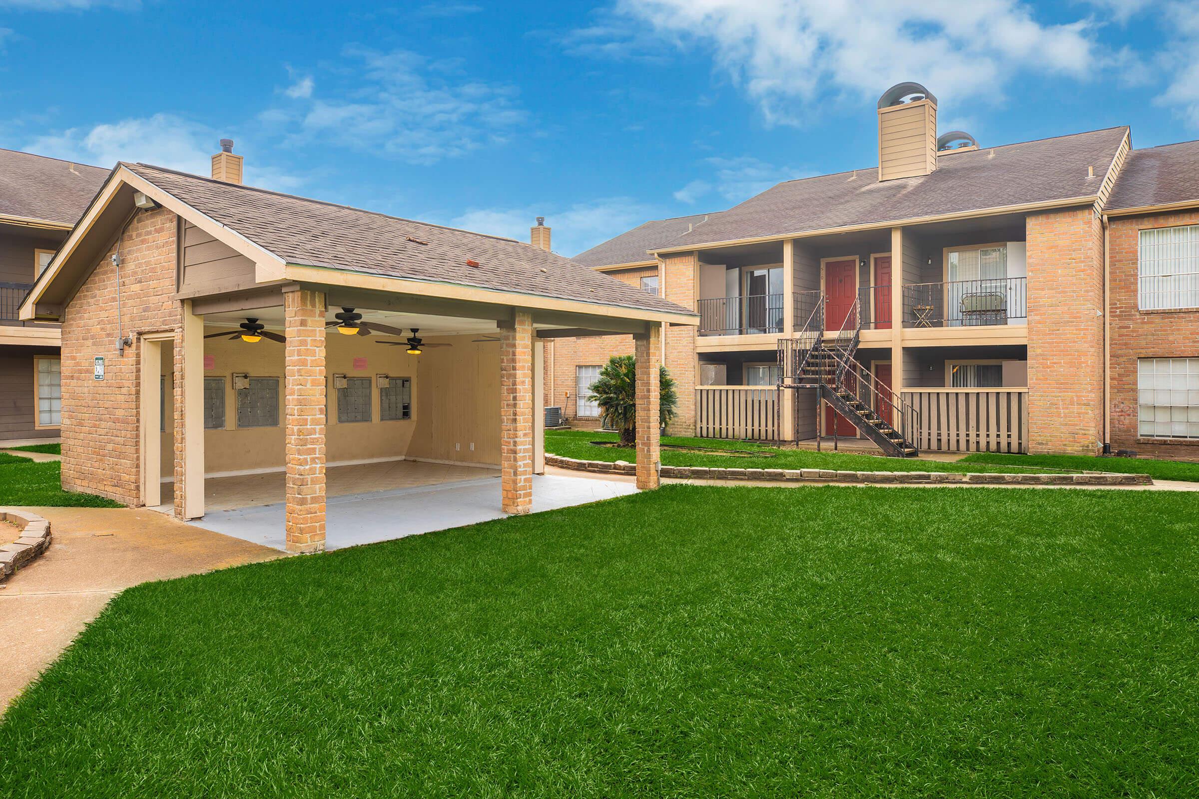 A spacious green lawn surrounded by residential buildings. In the foreground, there is a covered patio with ceiling fans, and in the background, a two-story apartment complex with a staircase leading to the upper level. The sky is clear and bright, enhancing the inviting atmosphere of the area.