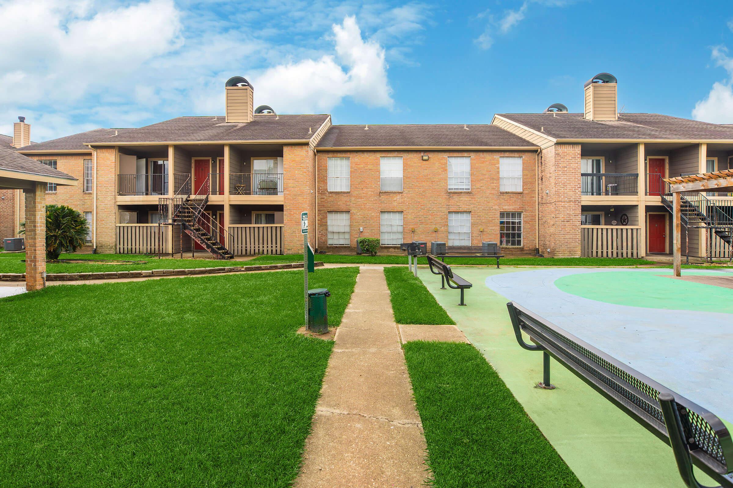 A well-maintained apartment complex with two-story brick buildings. Each building features balconies and staircases. In the foreground, a green lawn is bordered by a paved walkway leading to a playground area with a colorful surface and benches, set against a backdrop of a blue sky with scattered clouds.