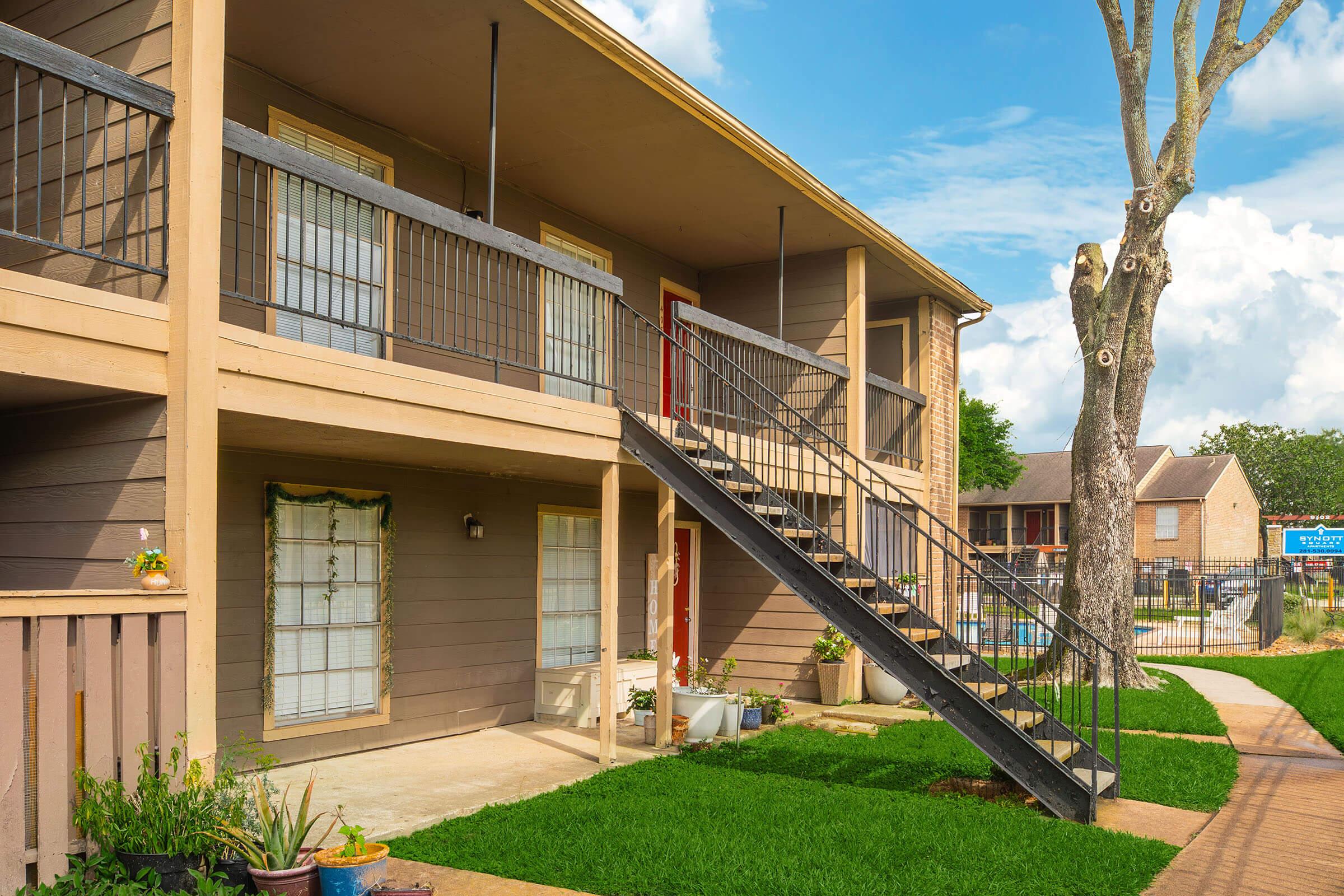 Two-story apartment building with a staircase leading to an upper balcony. The building features beige walls and red doors, surrounded by well-maintained grass and landscaping. In the background, a swimming pool is visible, along with other buildings. The sky is blue with a few clouds.
