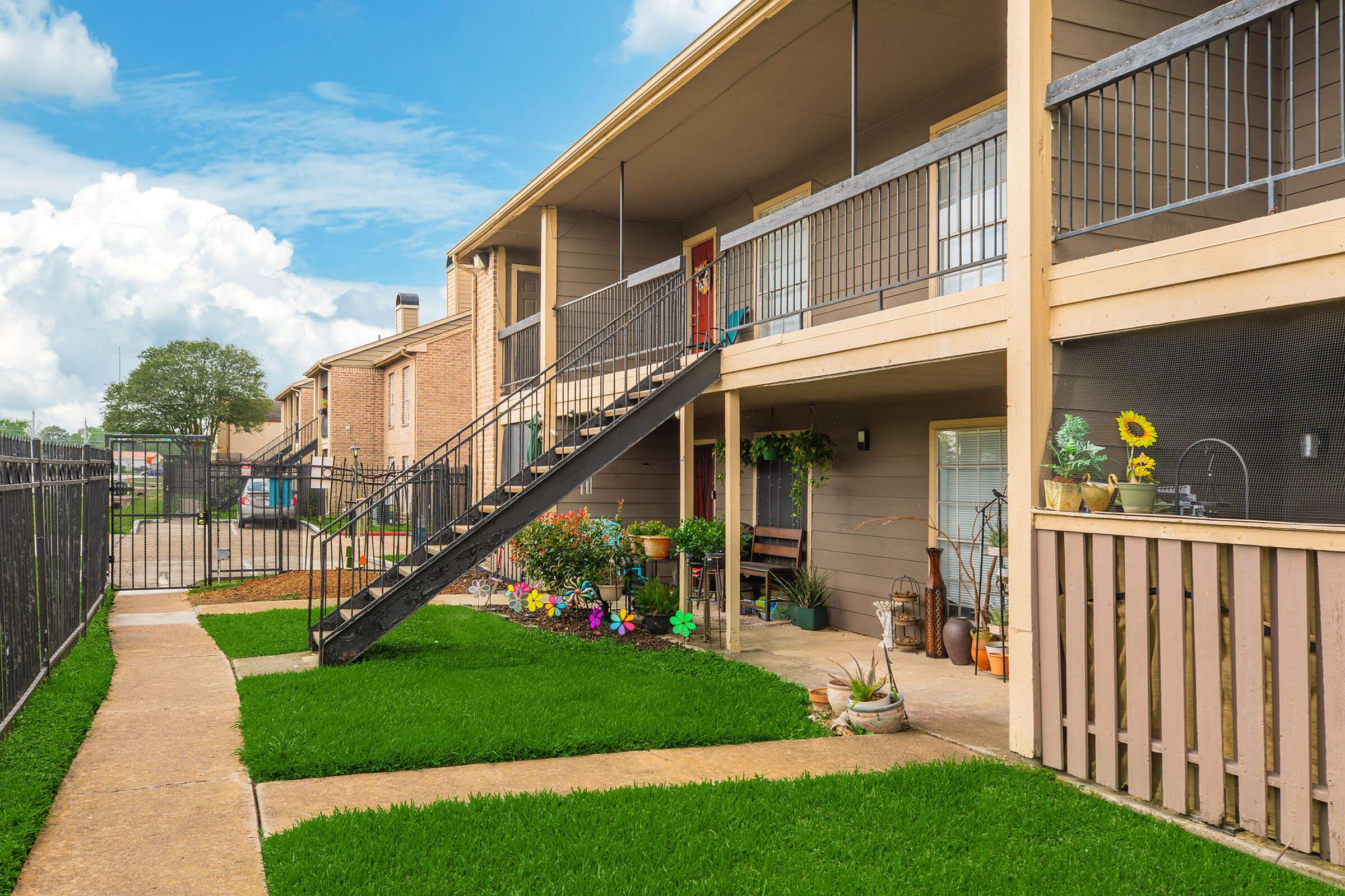 A well-maintained apartment complex featuring a two-story building with a staircase leading to the upper floor. The grassy area is neatly trimmed, and colorful flowers and potted plants are arranged along the porch and pathways. A fence surrounds the property, adding to its charm and privacy.