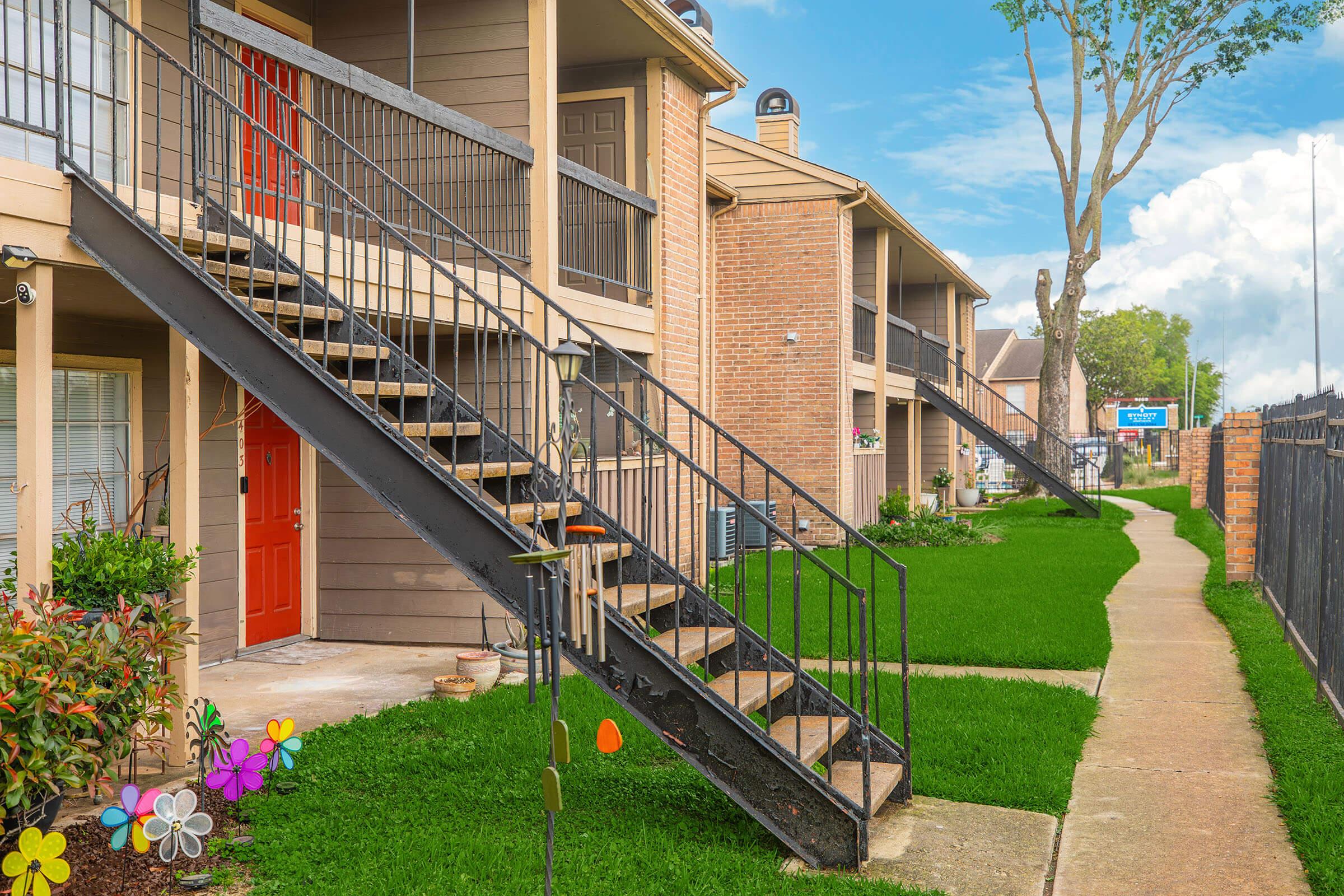 A view of a multi-story apartment complex featuring two staircases leading to upper units. The building has a mix of brick and siding, with vibrant plants and flowers in the foreground. A well-maintained pathway runs alongside the building, and there are blue skies in the background.